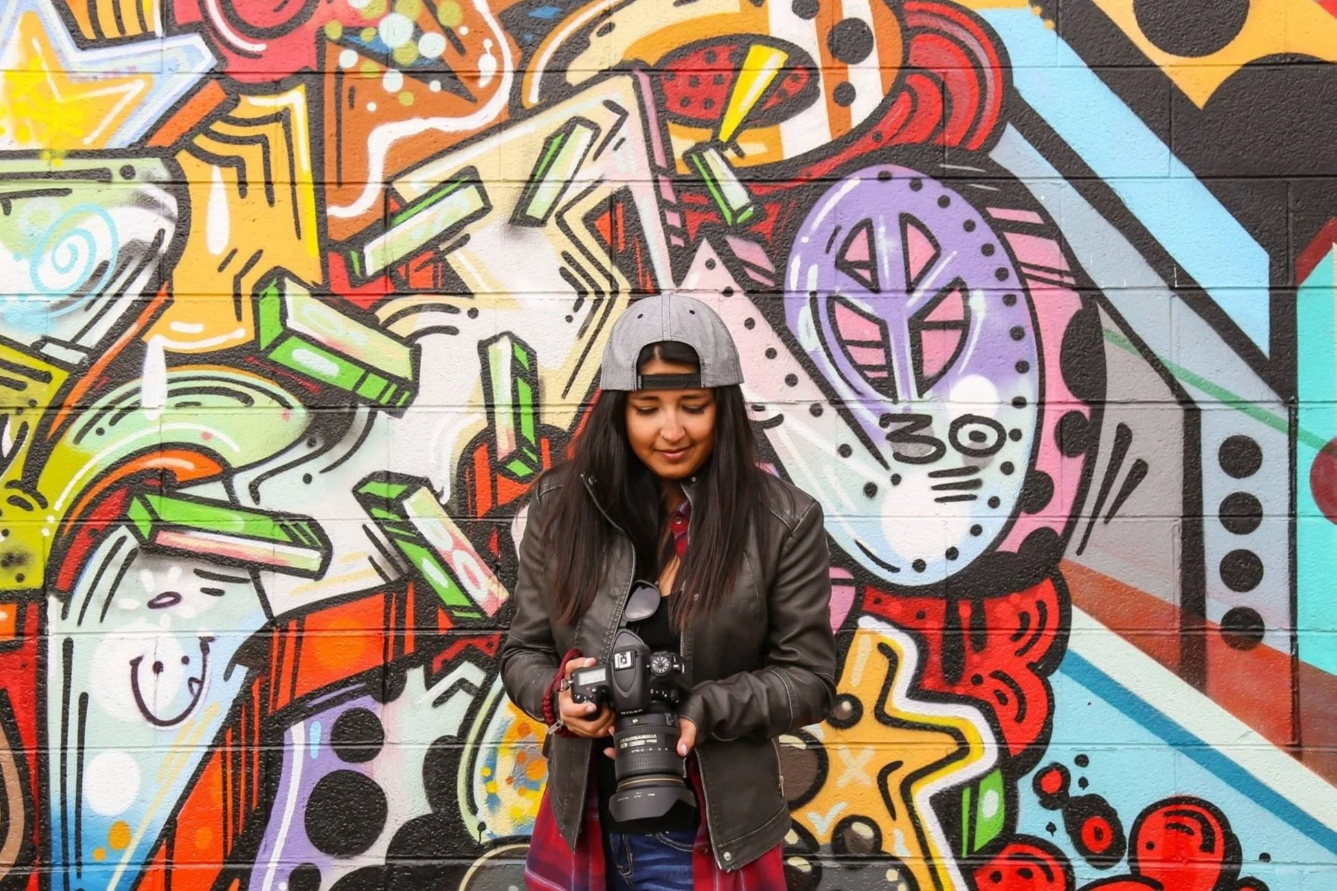 A woman with long dark hair wearing a gray backward cap, black leather jacket, and red plaid shirt is holding a professional camera with a zoom lens while standing in front of a colorful graffiti mural on a brick wall.