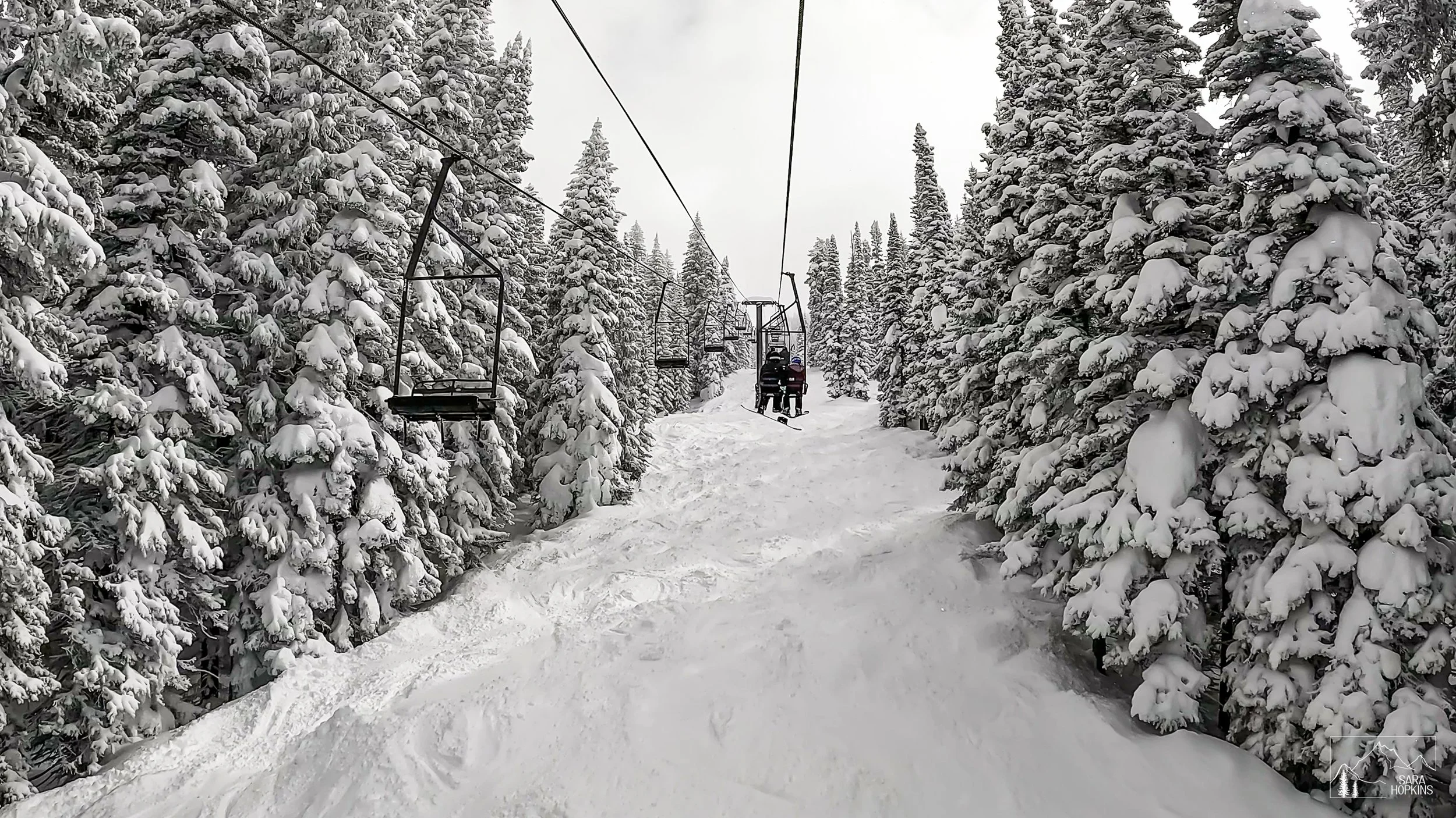 Snow-covered ski lift chairs on a cable above a snowy slope surrounded by snow-laden evergreen trees, with two skiers sitting on the lift.