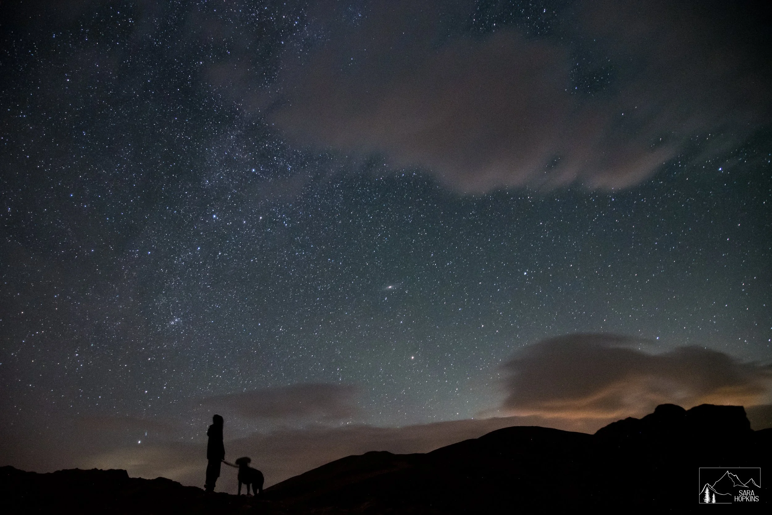Silhouette of a person and a dog standing on a hill at night, gazing at a clear starry sky with scattered clouds.