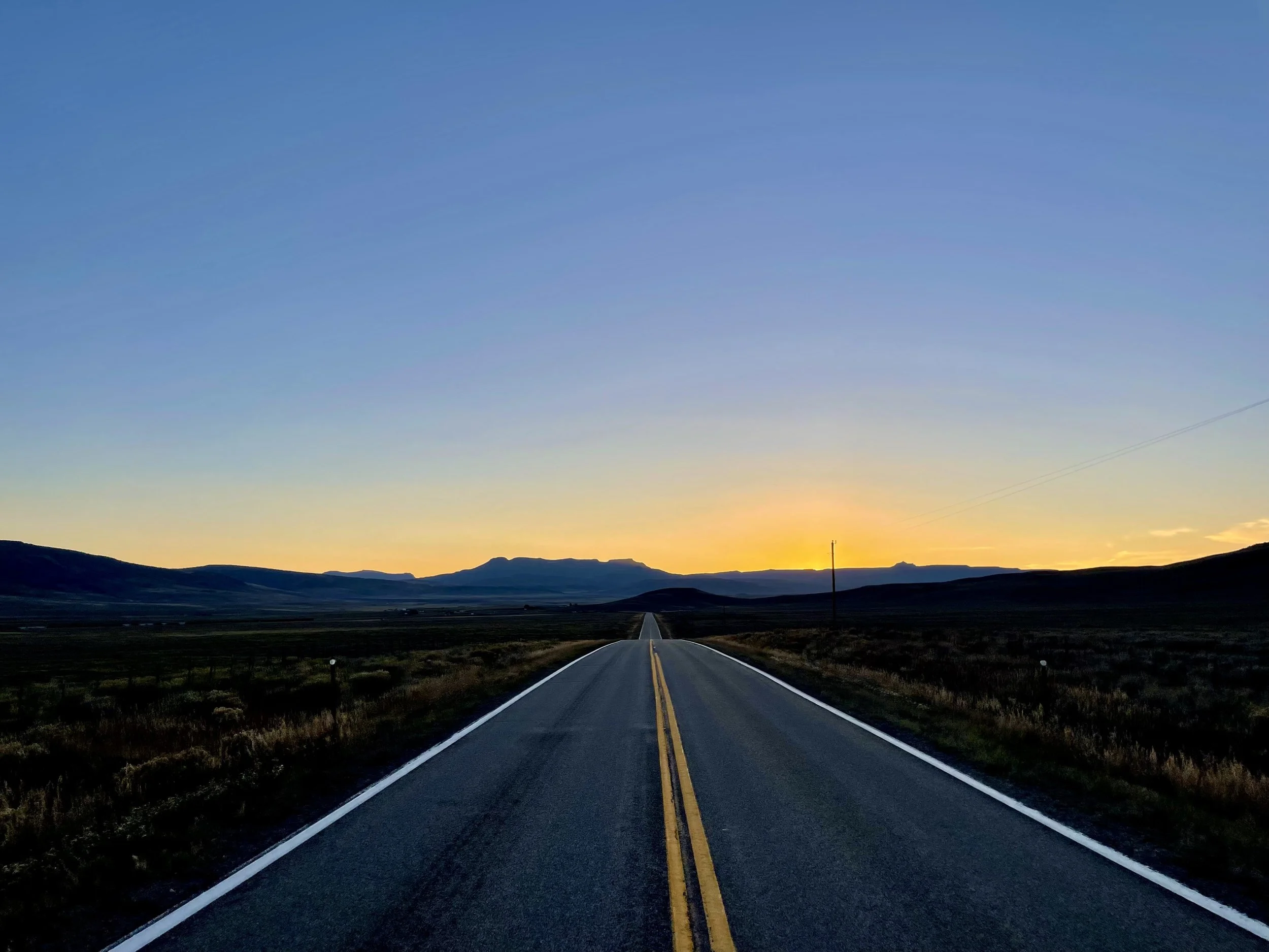A open, straight two-lane road in a rural area during sunset with mountains in the distance and a clear sky.