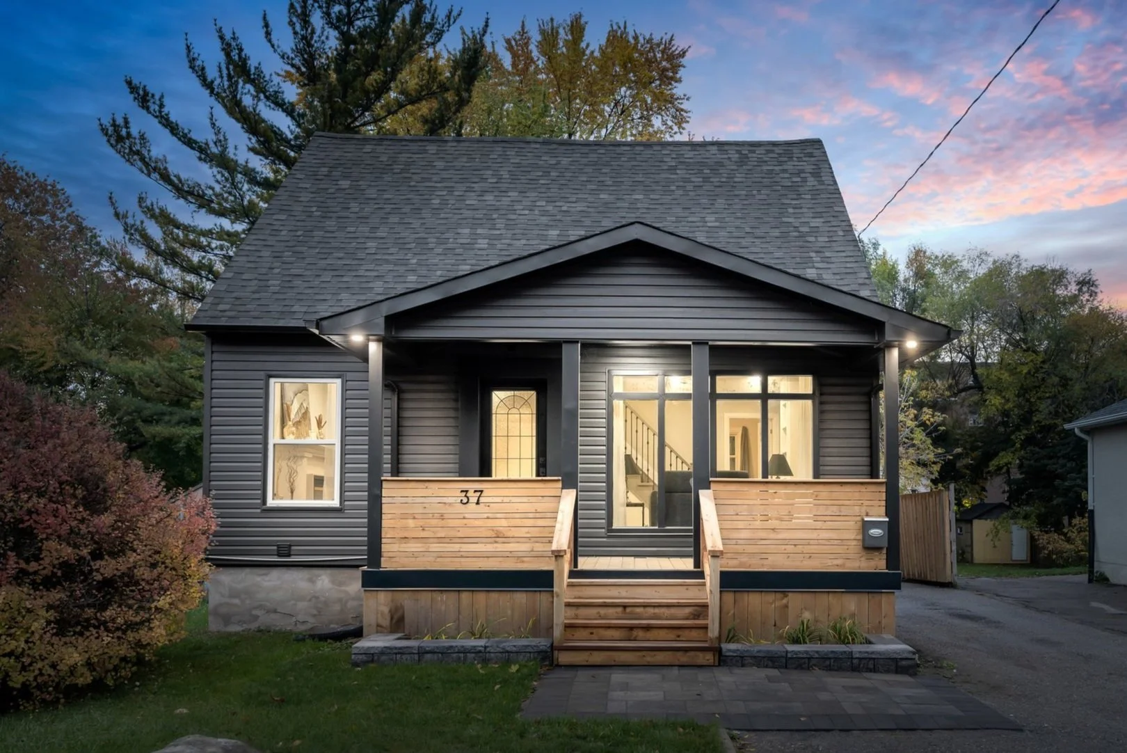 Small modern black house with wooden porch at dusk, surrounded by trees and illuminated by outdoor lights.