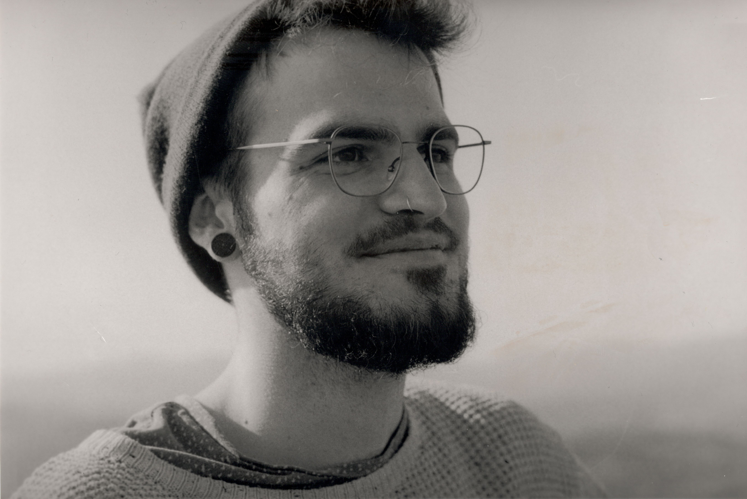 Black and white portrait of a bearded man wearing glasses, a beanie, and ear gauges, looking to the side with a slight smile.