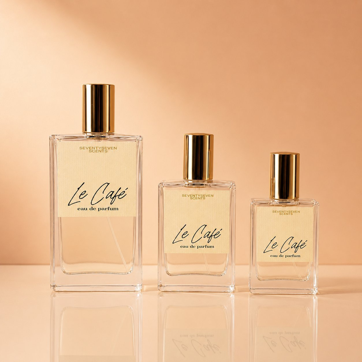 Le Café eau de parfum by SeventySeven Scents in three sizes, beautifully arranged on a warm peach reflective surface — a rich, indulgent handmade coffee gourmand from a UK indie fragrance house.