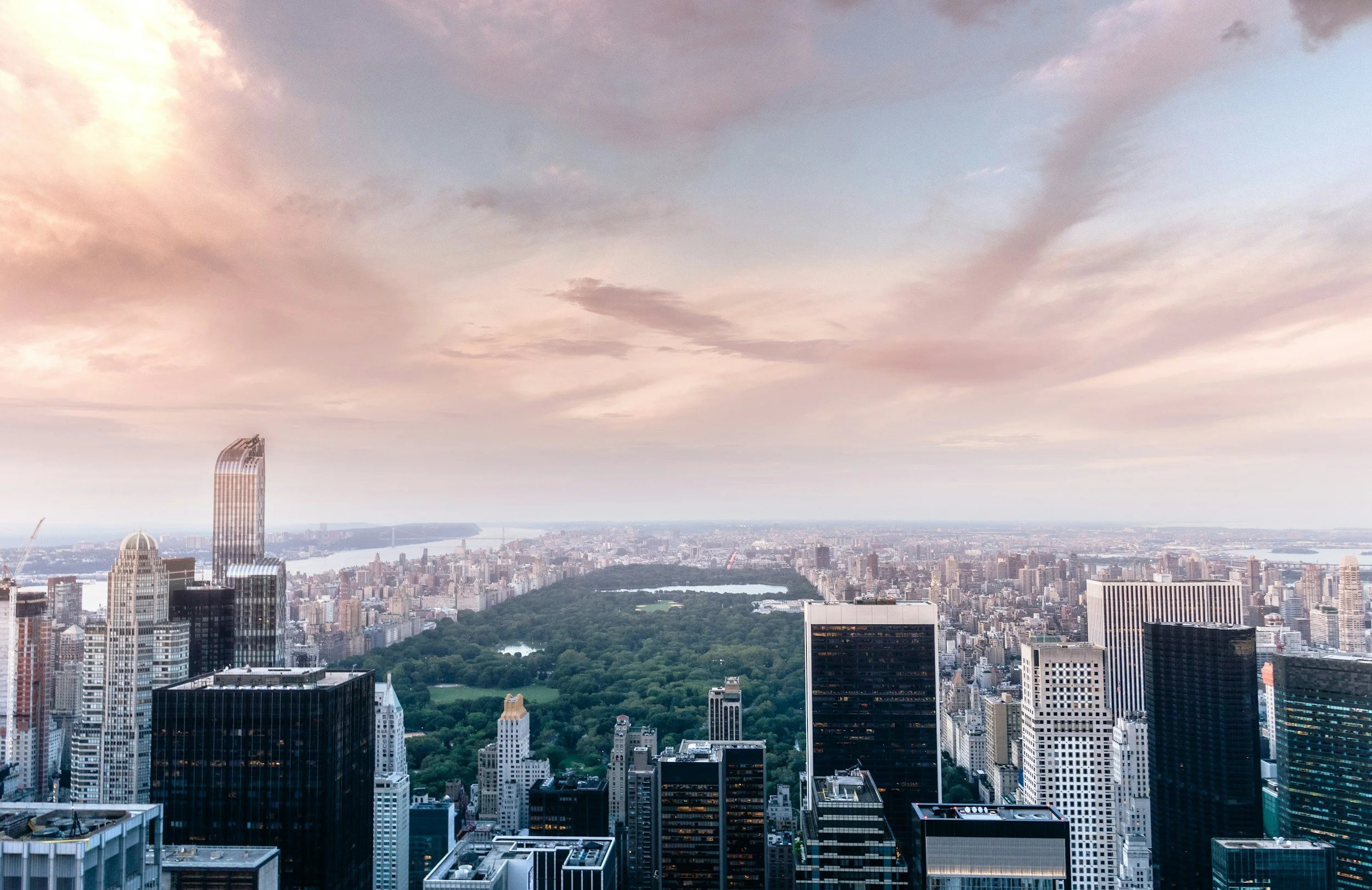 Aerial view of New York City skyline with Central Park in the center, skyscrapers, and a cloudy sky at sunset.