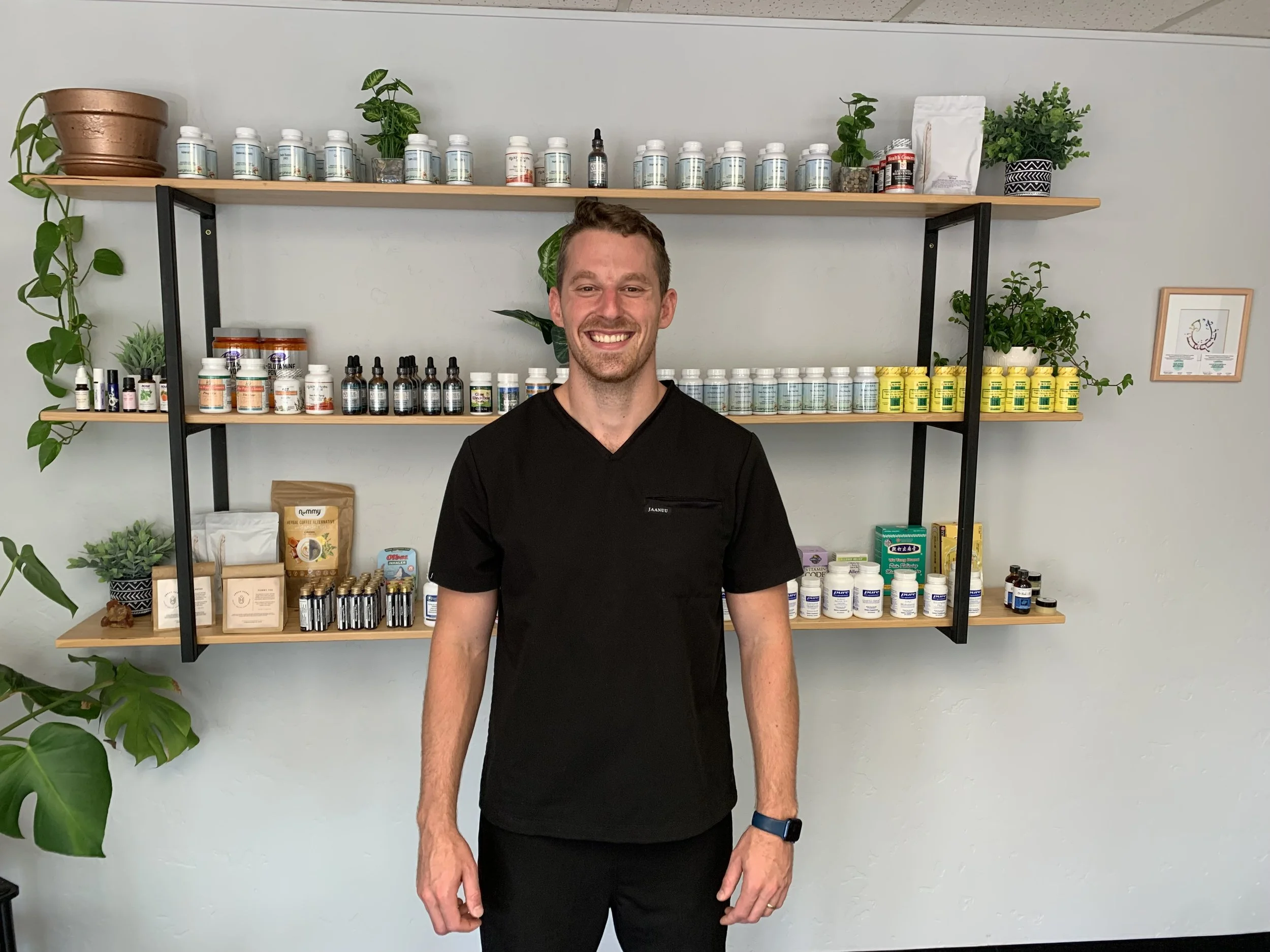 A man in black medical scrubs standing in front of a wall with shelves filled with supplement bottles, plants, and framed art, smiling at the camera.