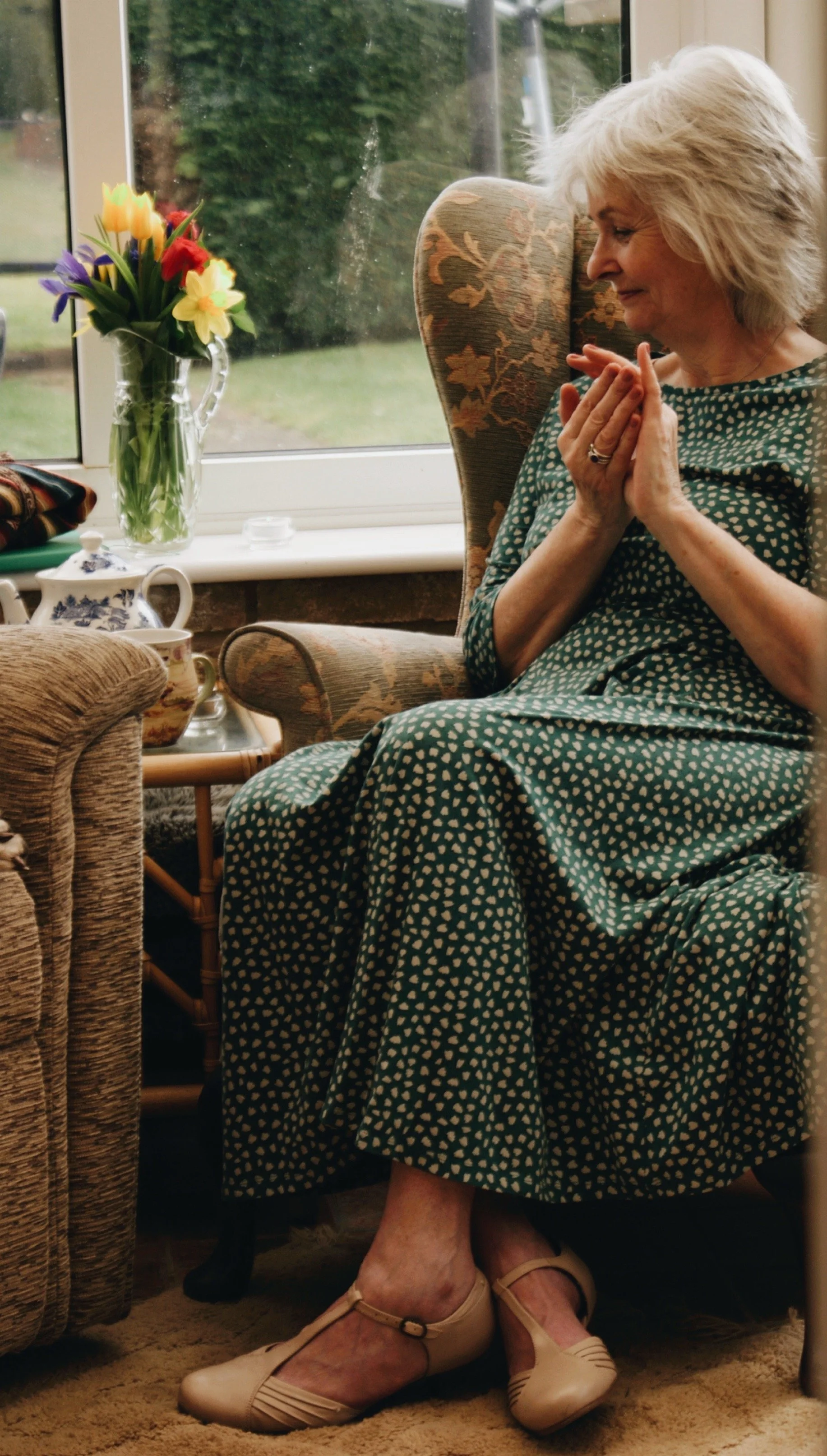 Annie Barber sitting in conservatory in emerald green dress, she is holding a crystal in her hands and is looking downwards. There is a vase of spring flowers in the background.