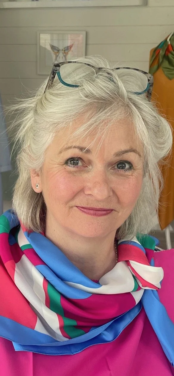 Headshot image of Annie Barber looking towards the camera, she is waring a scarf of pinks ad blues and has her glasses on top of her head. She has a gentle smile on her face.