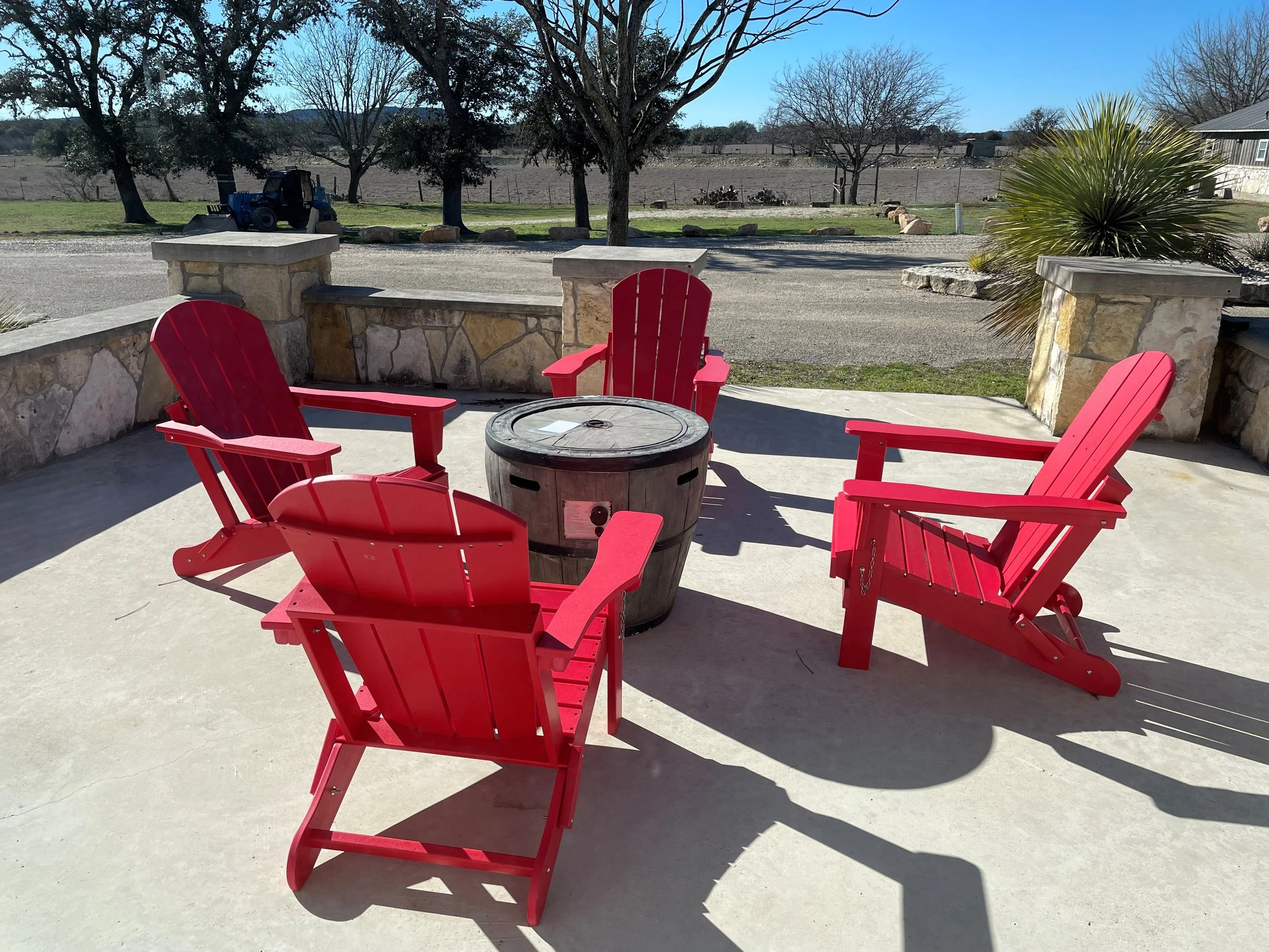 Four red Adirondack chairs around a small round table with a hole in the center, on a concrete patio. Behind the patio is a stone wall, trees, and a grassy field with a tractor parked on the grass. The sky is clear and blue.