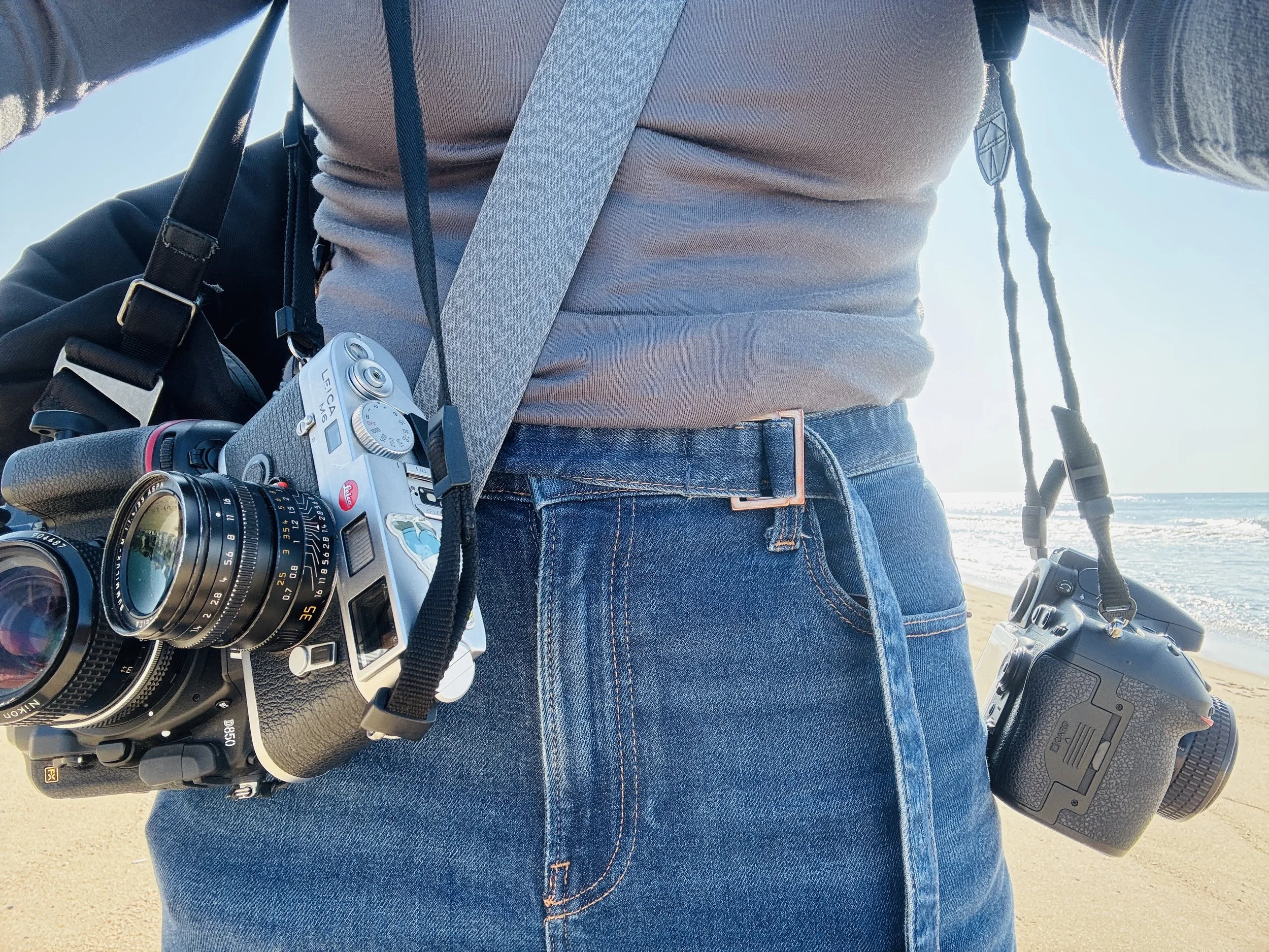 Angela Cappetta is a New York–based wedding and portrait photographer. Person standing on the beach with two cameras hanging from their waist, one around their neck and the other attached to their belt, with the ocean in the background.