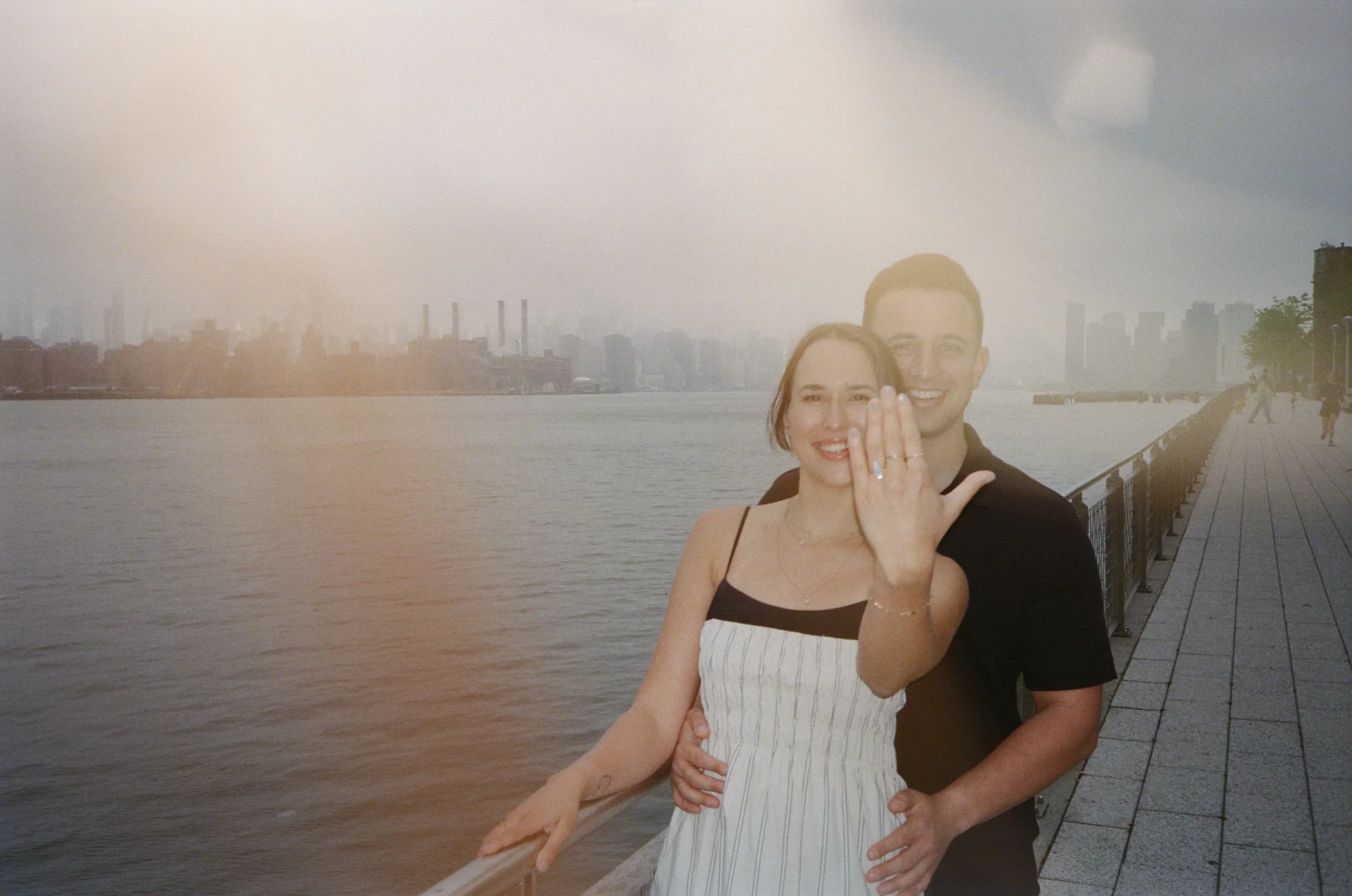 Moody film photo of a couple, showcasing an engagement ring, during a romantic engagement session at Domino Park in Brooklyn.