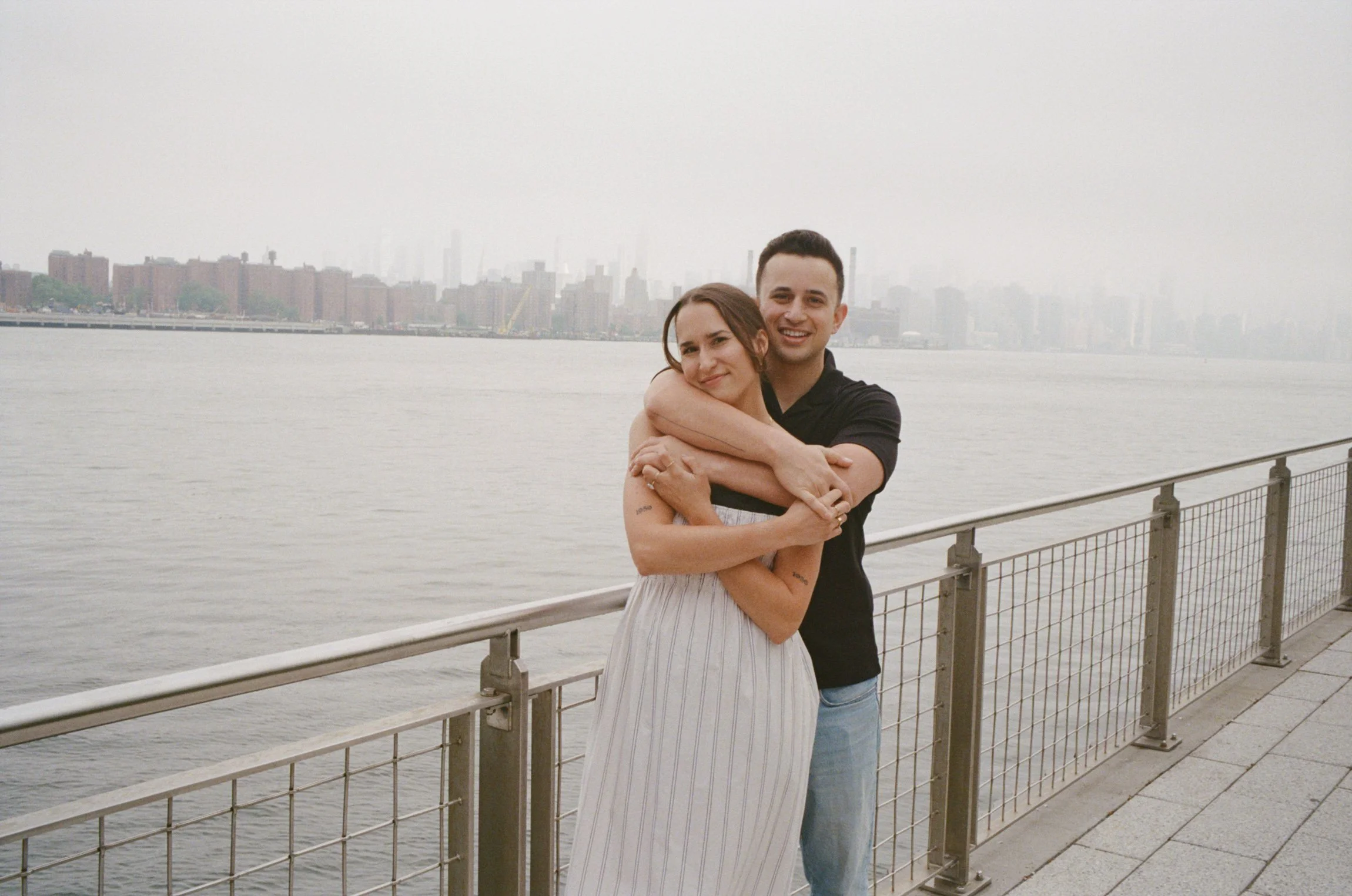 Couple showing off her ring during a romantic film photography engagement shoot at Domino Park in Brooklyn, with the Manhattan skyline and Williamsburg waterfront in the background.