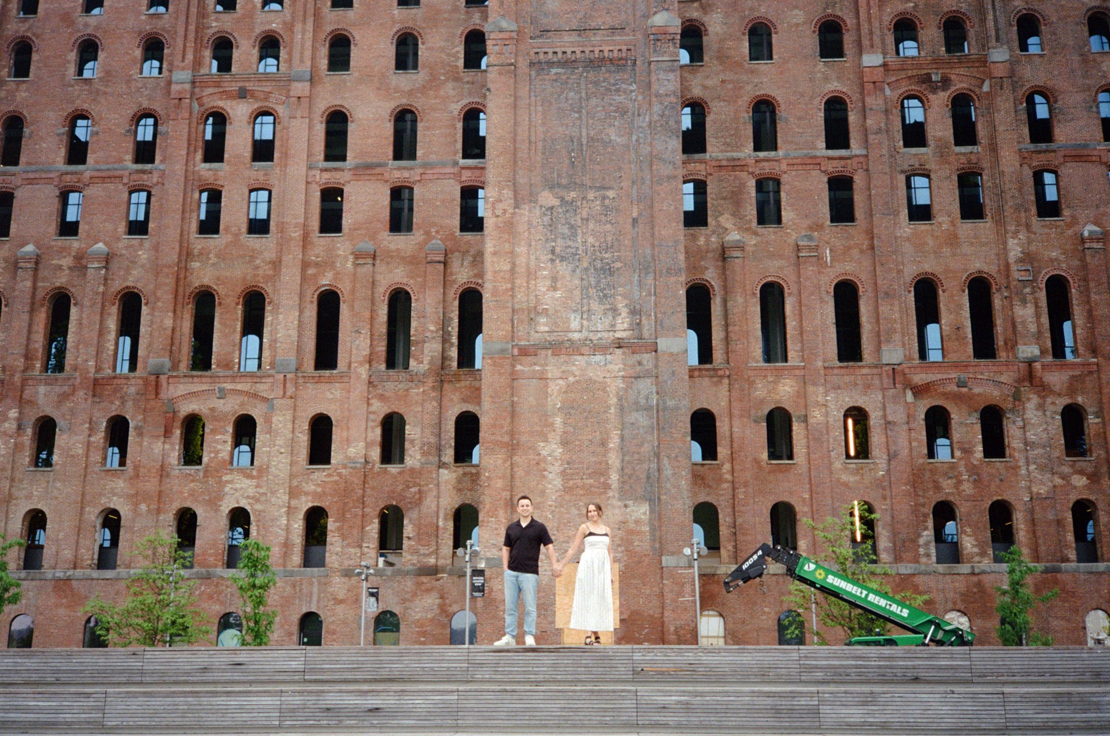 Film photo of a couple in front of the Domino Sugar factory during a romantic engagement session at Domino Park in Brooklyn.