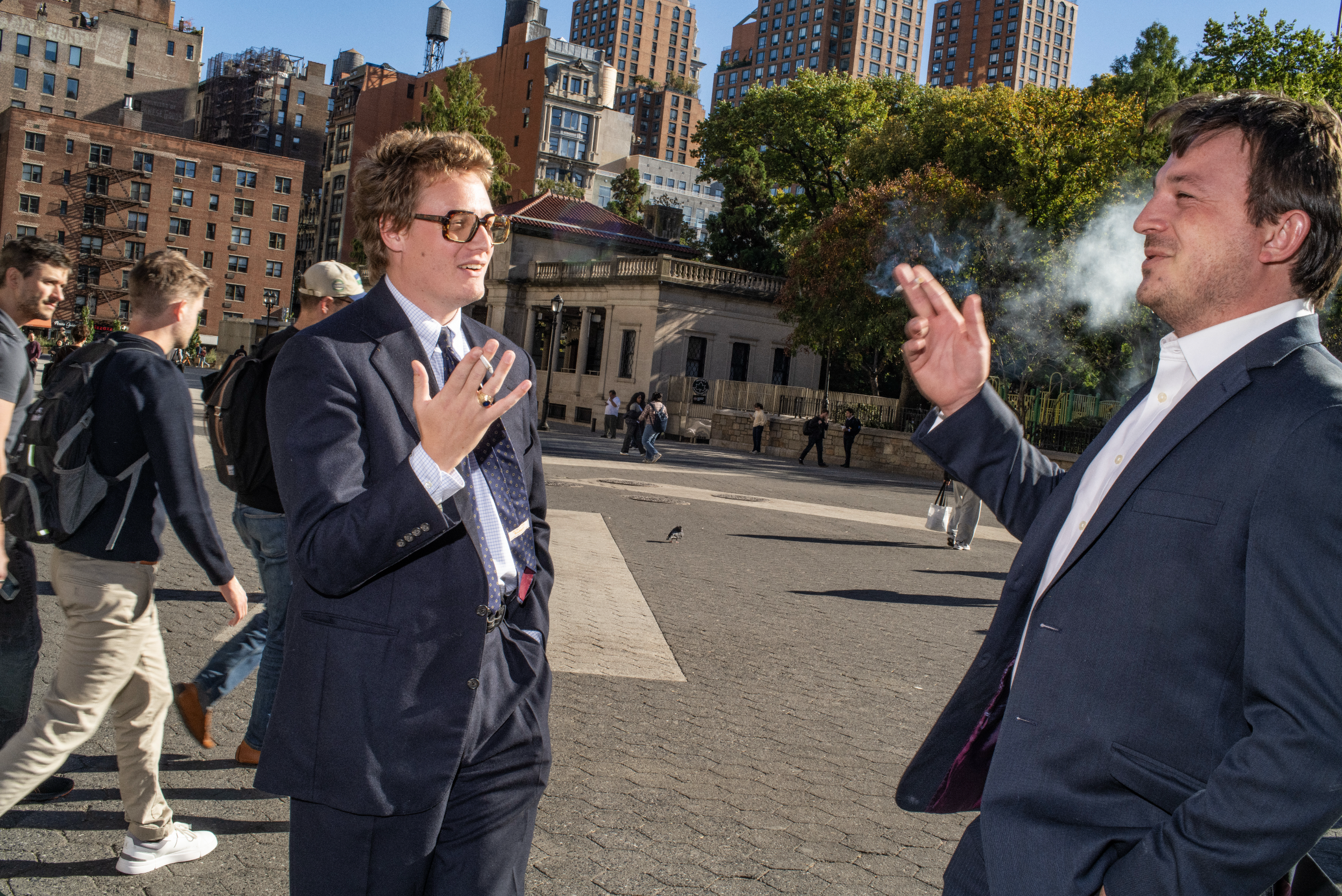 Two young men in blue suits smoking in a park.