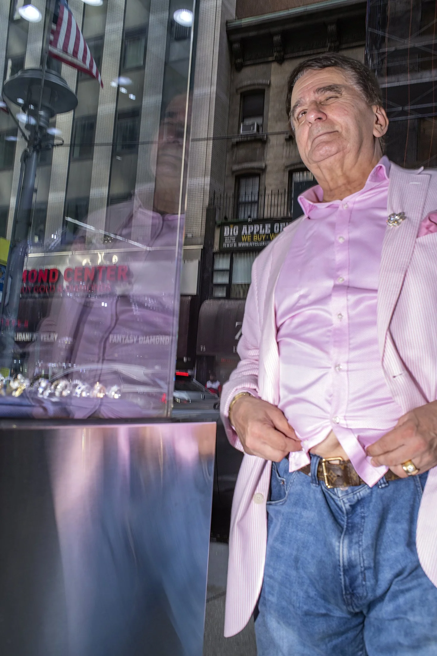 A man in a pink shrt stands beside his reflection in a shop window on 47th Street