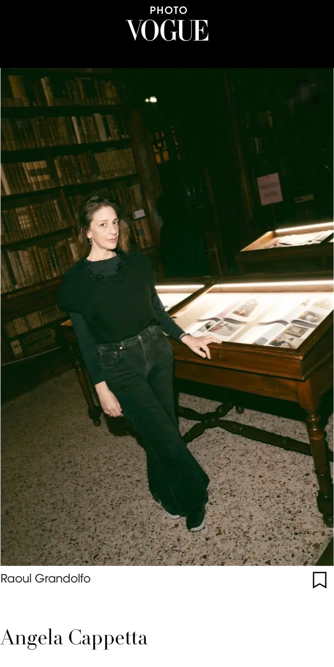 A woman with brown hair and fair skin wearing a black top and black jeans, standing beside a lit display case in a dimly lit library or museum with dark wooden shelves filled with books in the background.