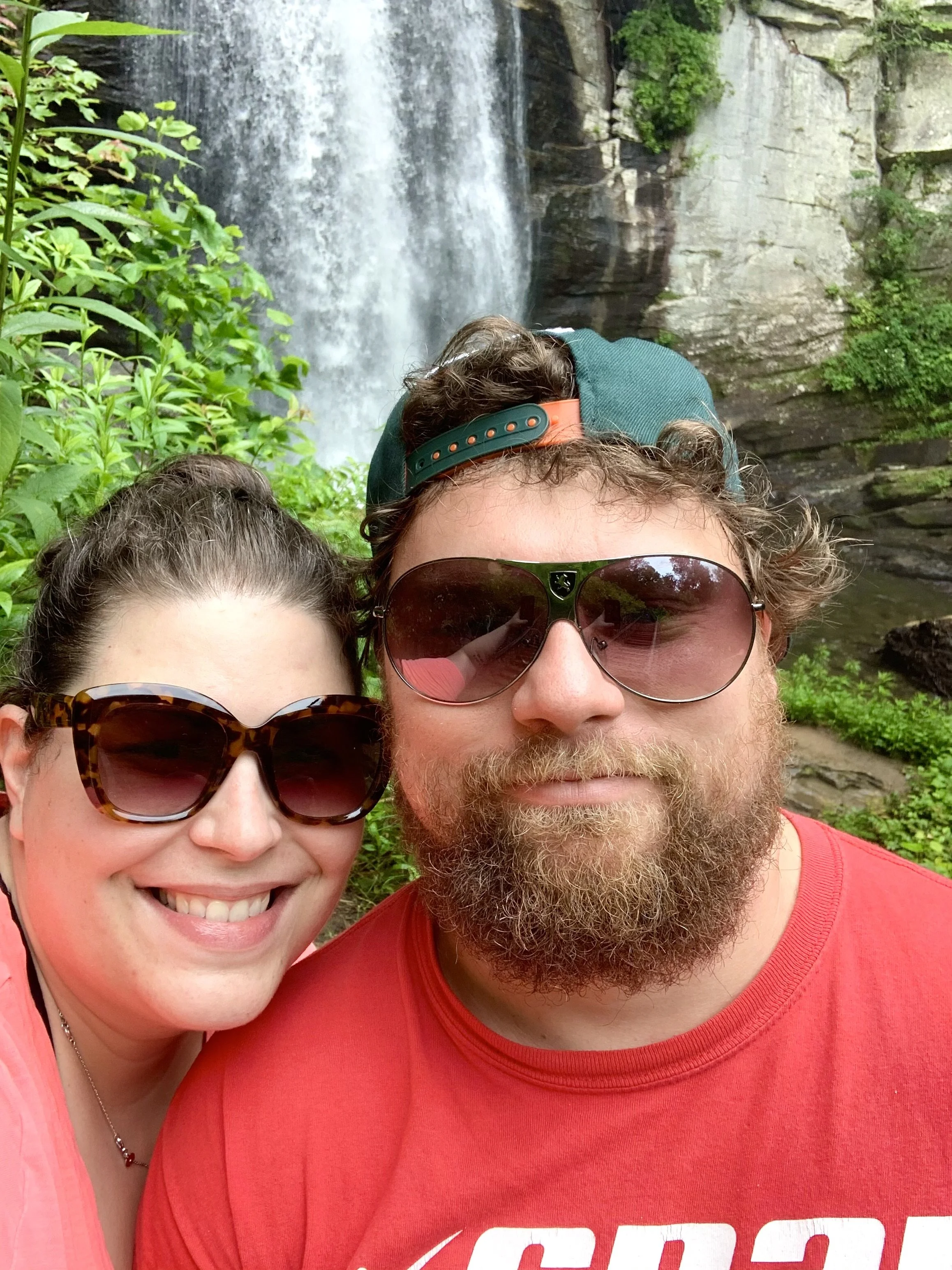 Laura and Adrian are smiling in front of a waterfall they hiked to.