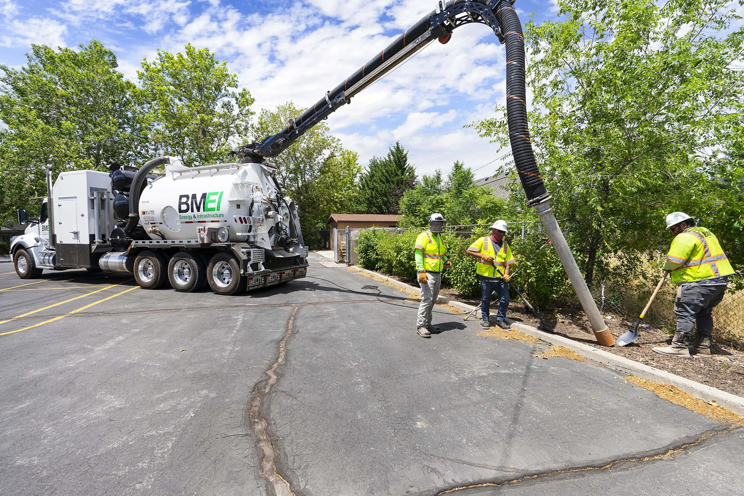 Workers in safety vests and helmets repairing a fallen utility pole with a vacuum truck nearby in a parking lot.