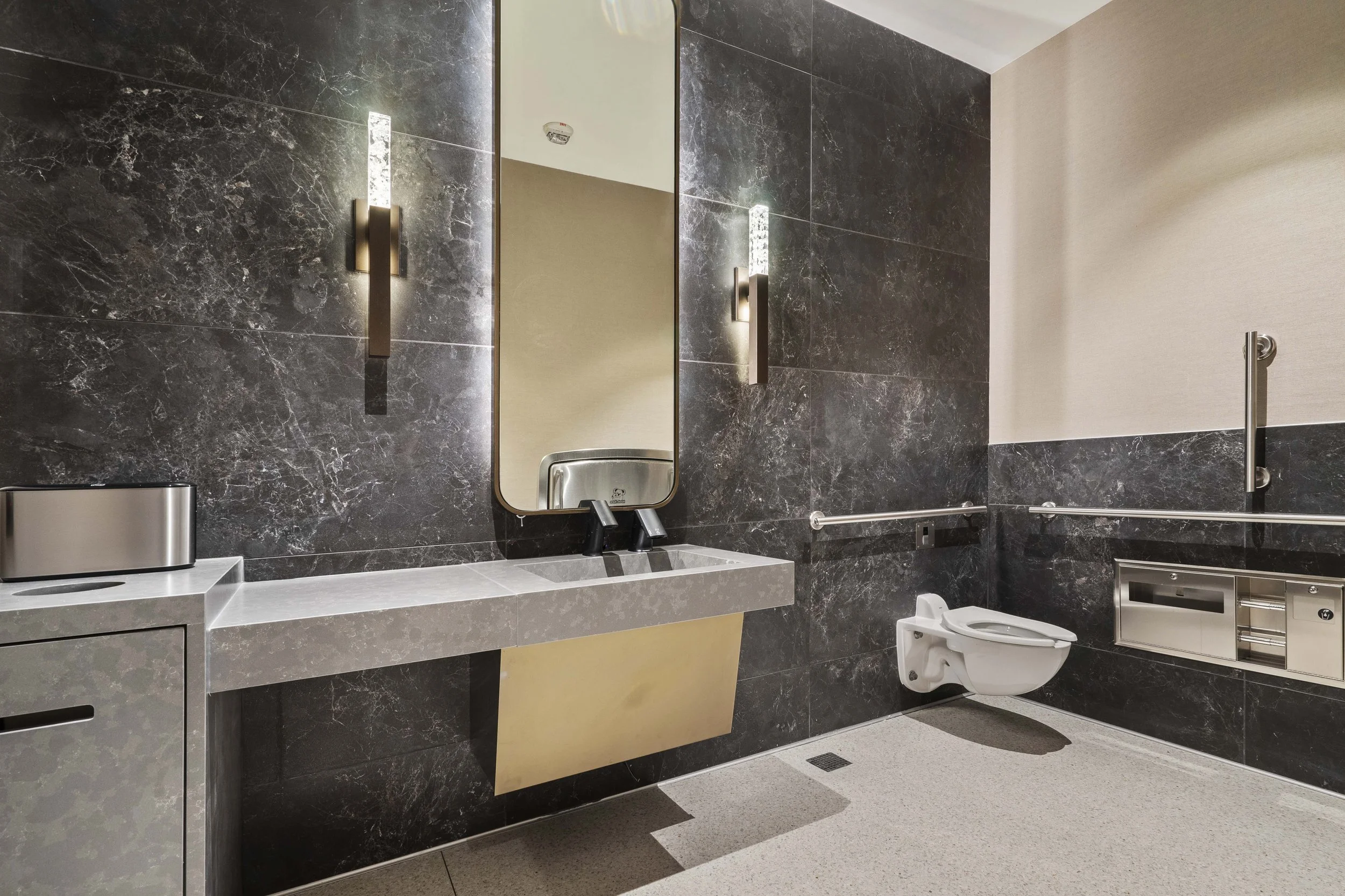 Modern accessible restroom with black marble walls, a large mirror, wall sconces, and a toilet with grab bars.
