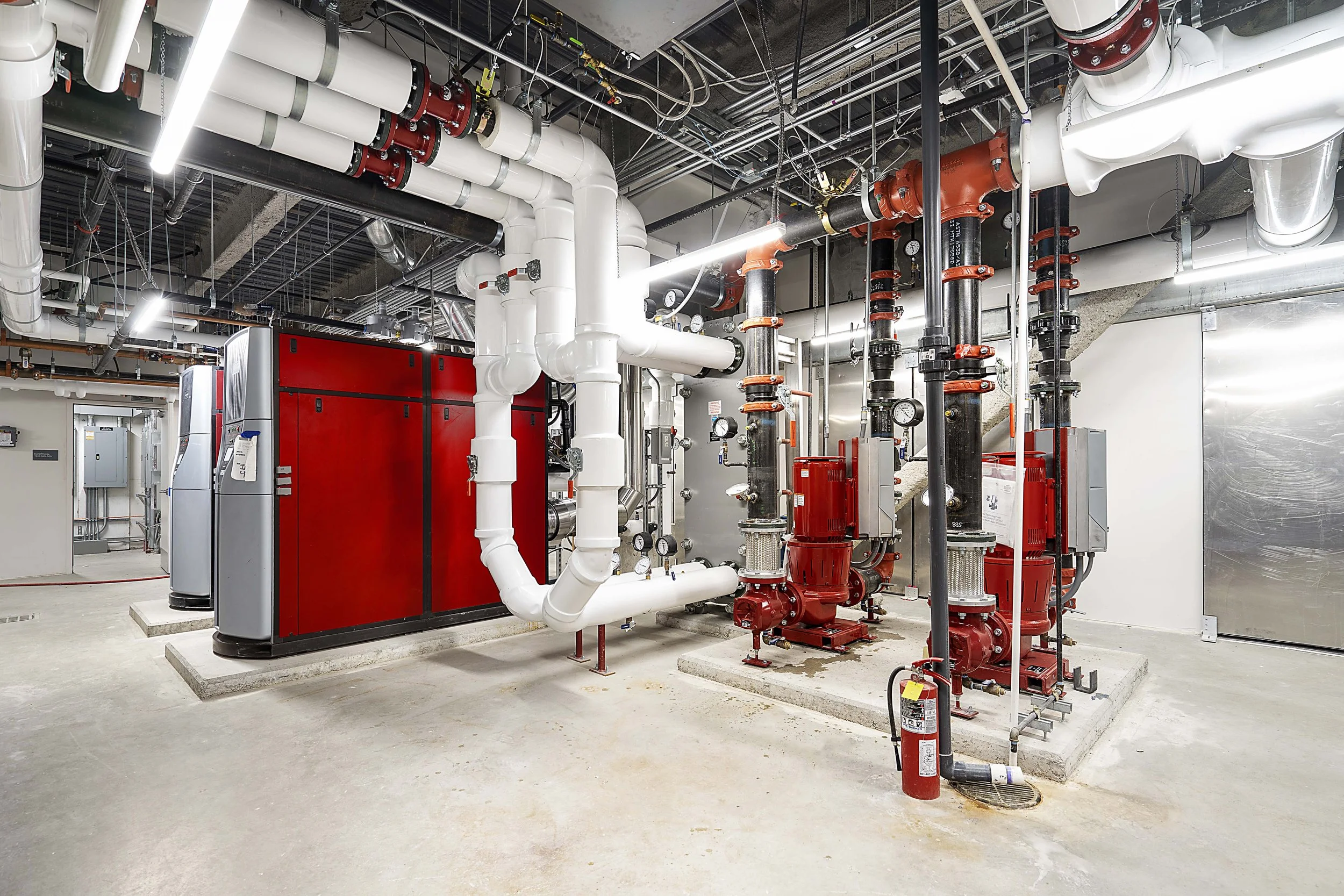 Industrial mechanical room with large red and gray machinery, white and black pipes, gauges, and fire extinguisher on concrete floor.