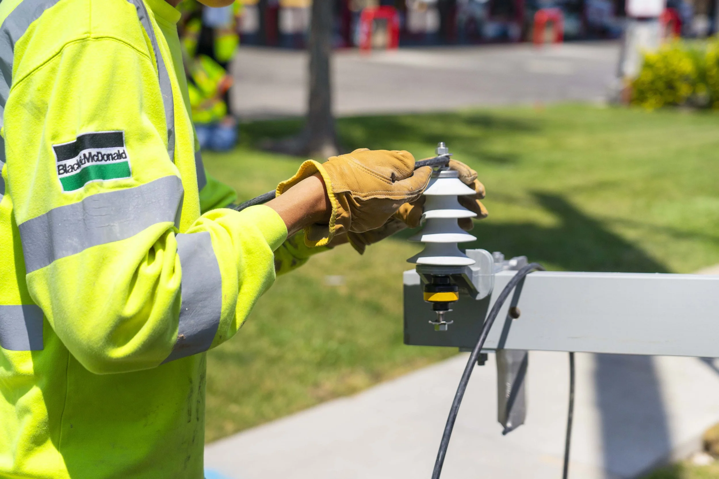 A worker wearing a high-visibility yellow jacket with a Black McDonald patch, gloves, and safety gear is handling electrical equipment outdoors during the daytime.
