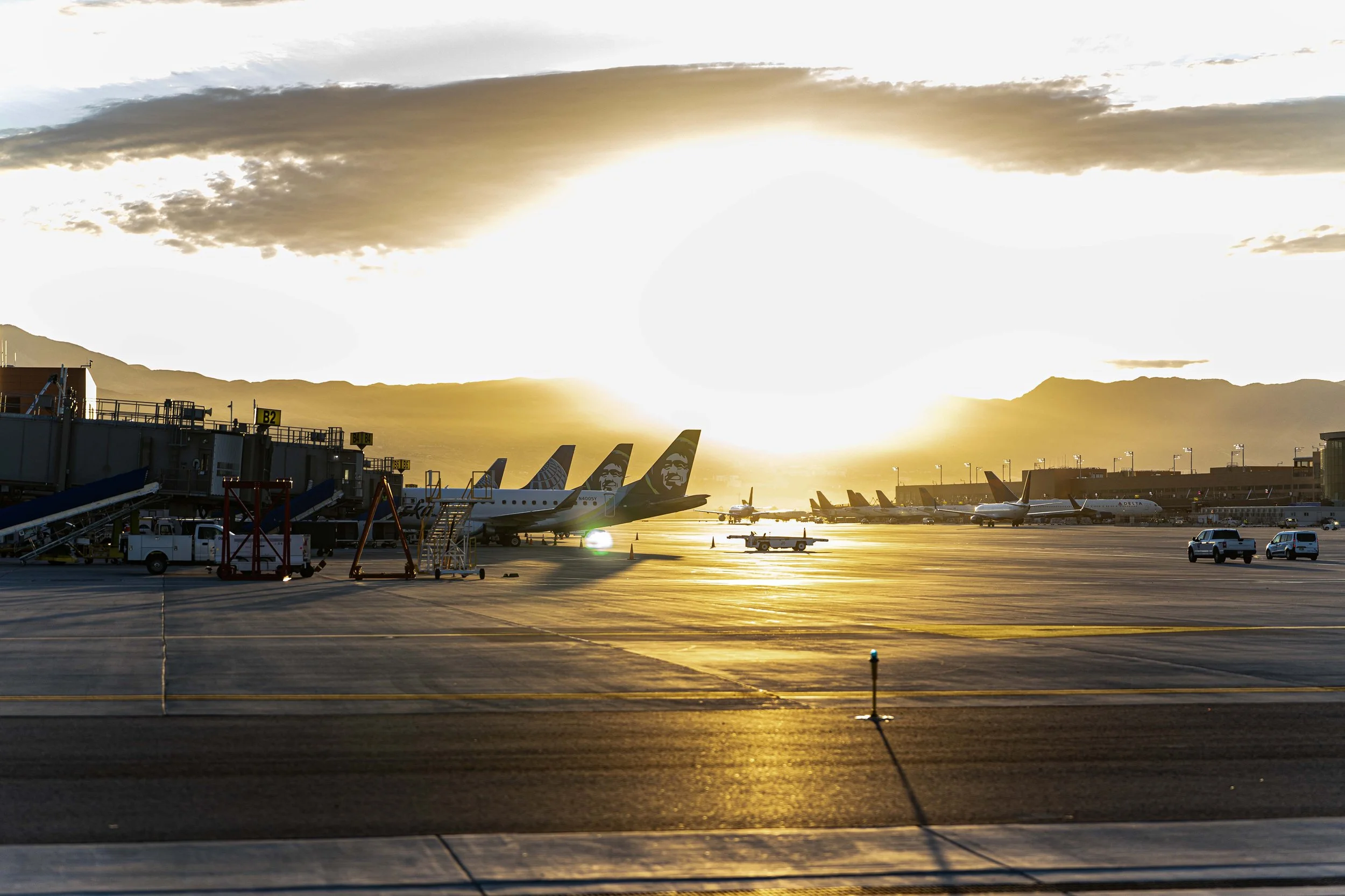 View of an airport tarmac at sunset, with airplanes parked at gates, some branded with Alaska Airlines and Delta, and vehicles moving around. Mountain range in the background, with clouds overhead.