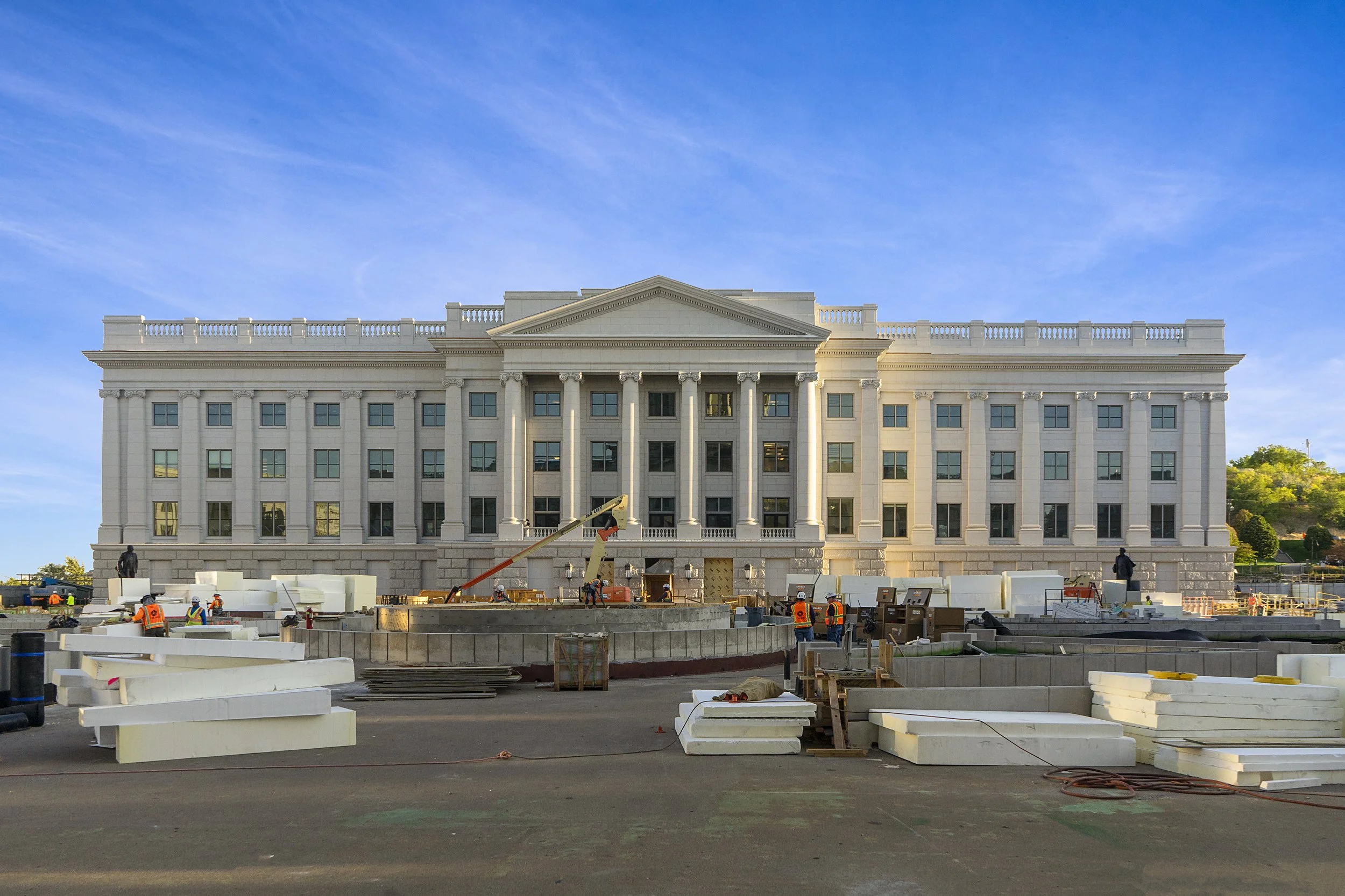 Construction workers building a large, white, neoclassical style government or institutional building with columns and a pediment, on a clear day.
