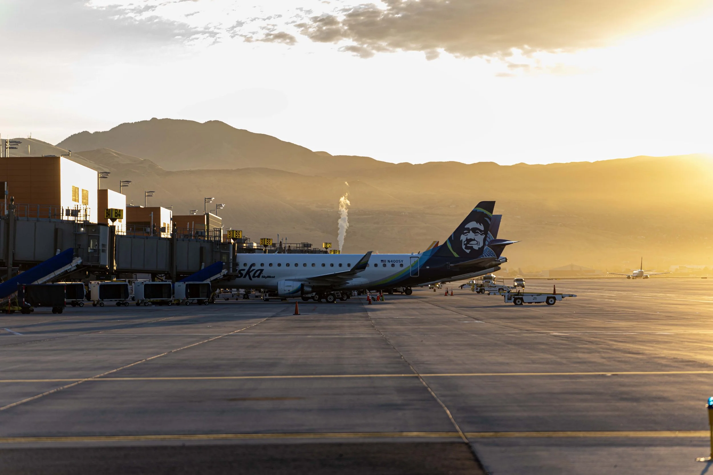 An airplane at an airport gate during sunset, with mountains in the background and a small plane on the runway.