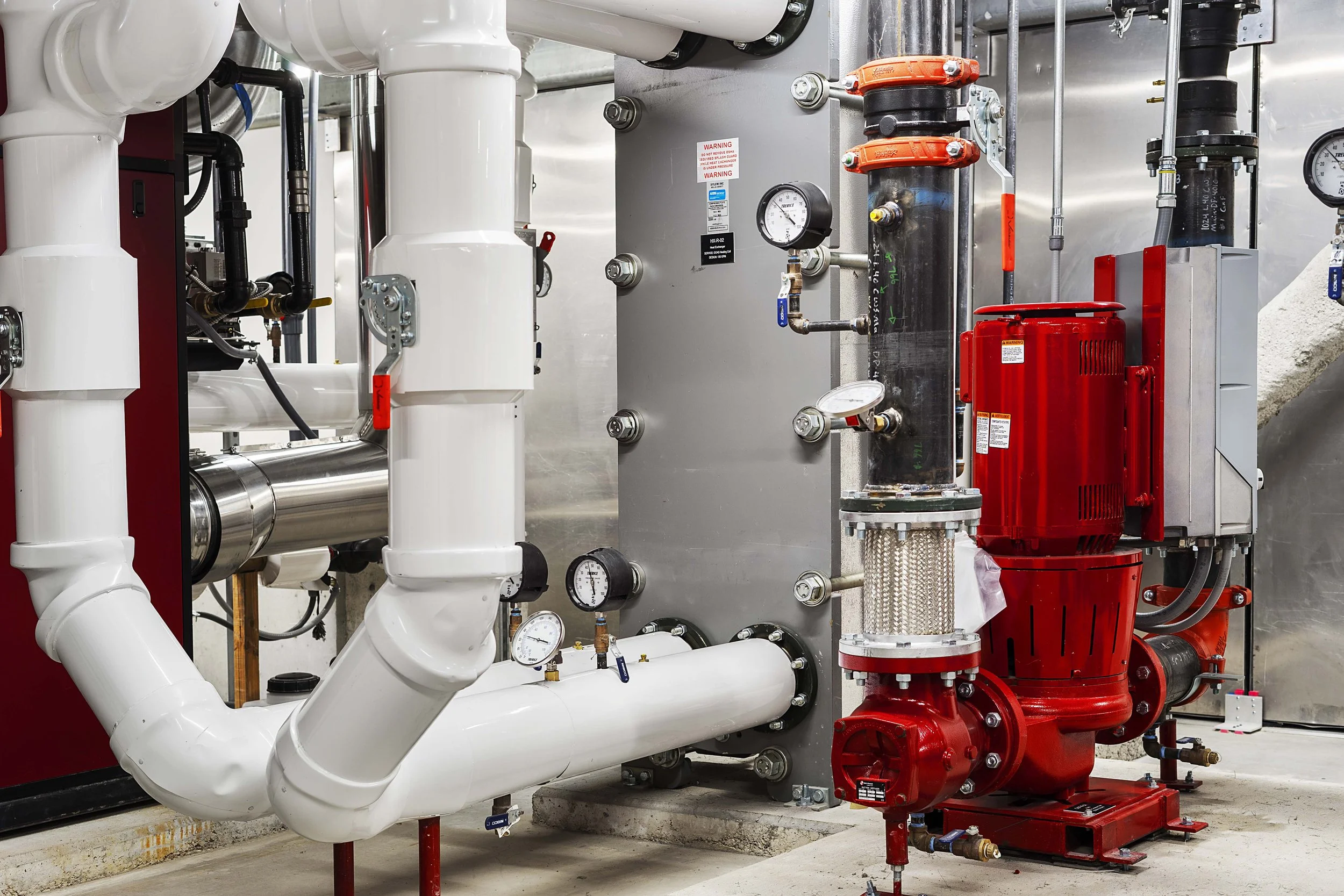 Industrial mechanical room with white pipes, gauges, and a red pump.