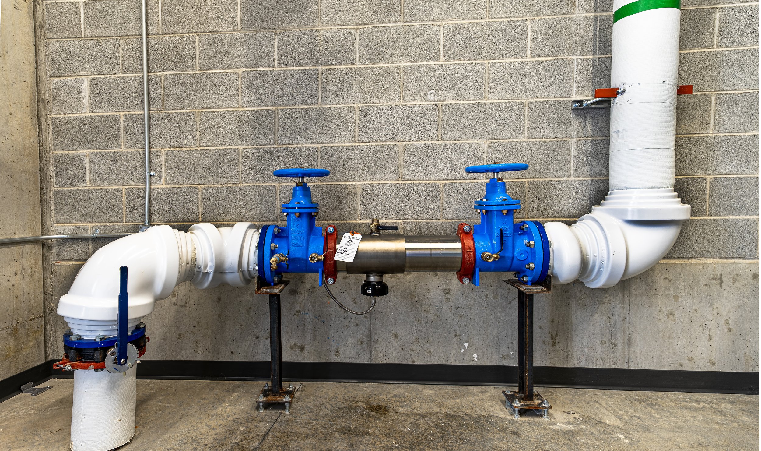 Industrial water piping system with white pipes, blue valves, and red fittings against a cinder block wall.