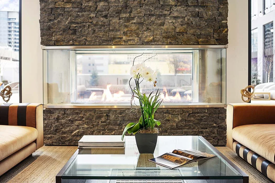 Modern living room with two beige sofas, a glass coffee table, a potted orchid, and a wall with a built-in fireplace and a large window offering a city view.