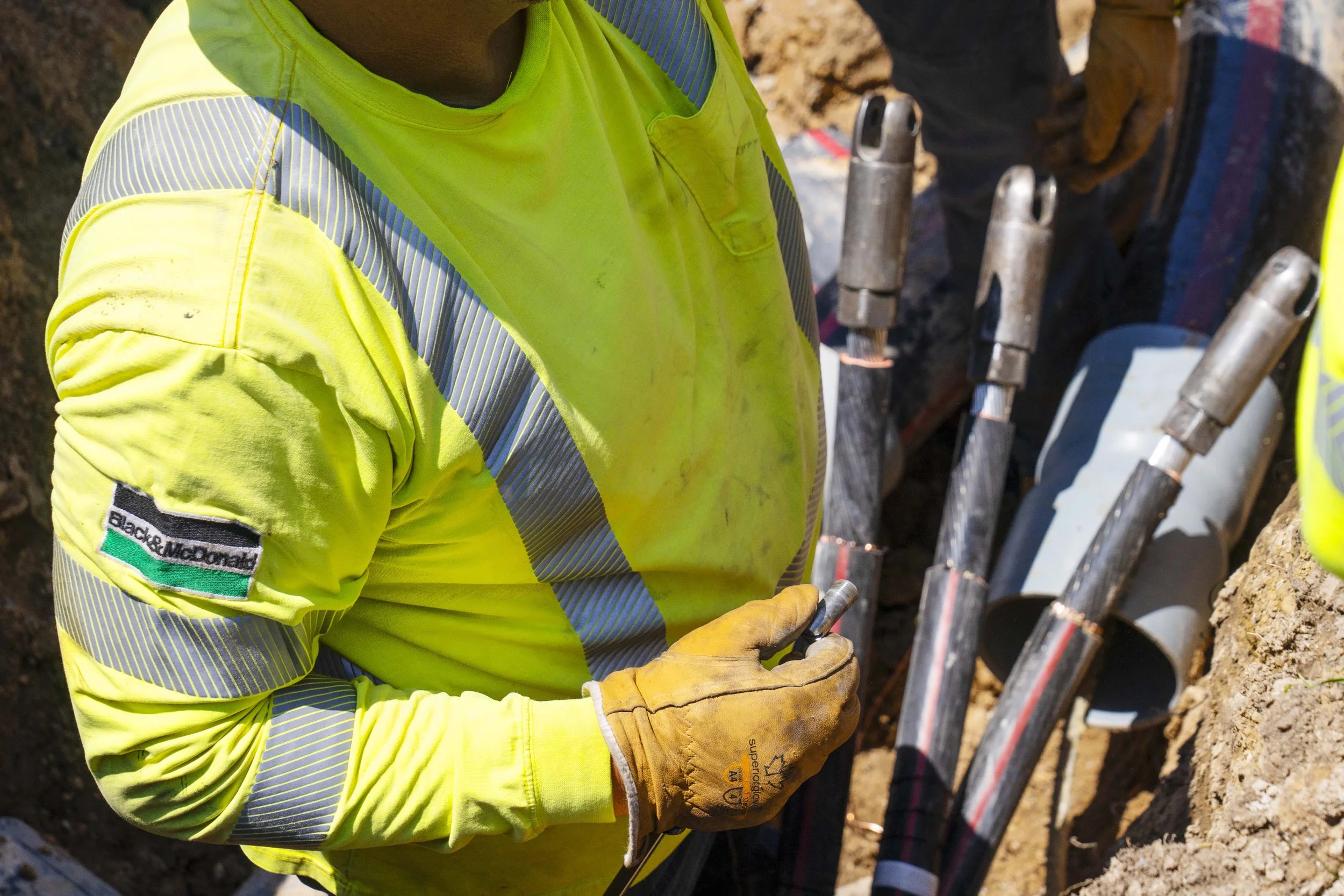 A construction worker wearing a high-visibility yellow safety shirt with reflective stripes, holding a camera or phone, and surrounded by pipes and equipment at a construction site.