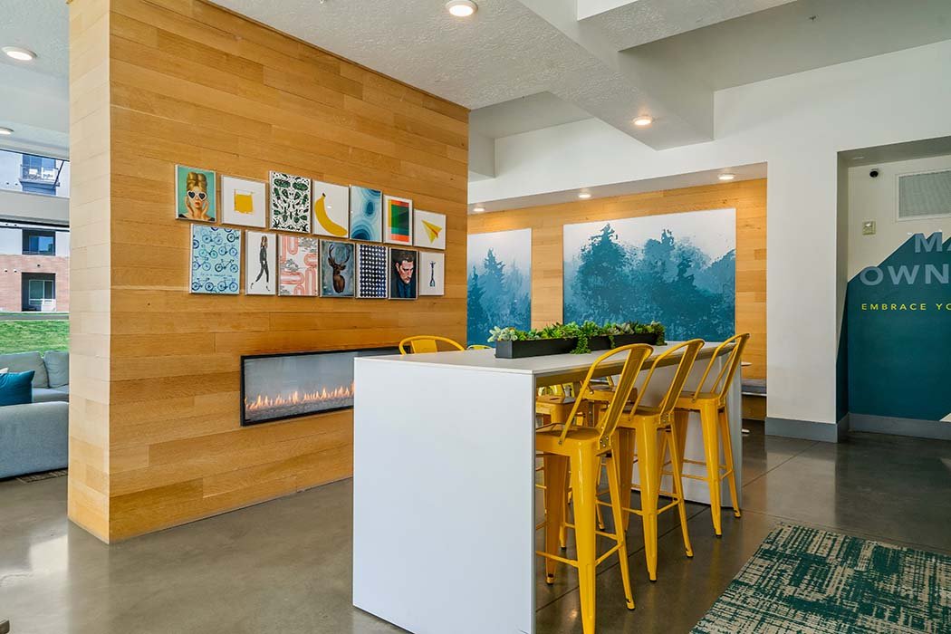 Modern lounge with yellow chairs around a white high table, wooden wall with colorful artwork, a long rectangular fireplace, and wall art of blue trees in the background.