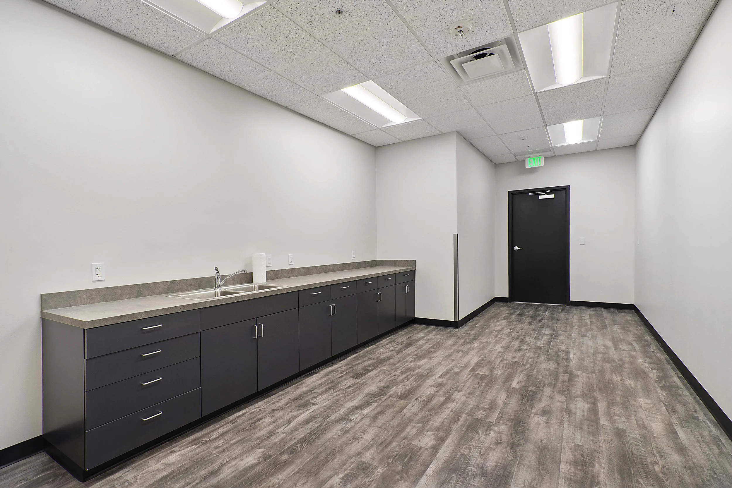 Empty room with a counter, sink, kitchen paper, and multiple electrical outlets, with a black door and illuminated exit sign