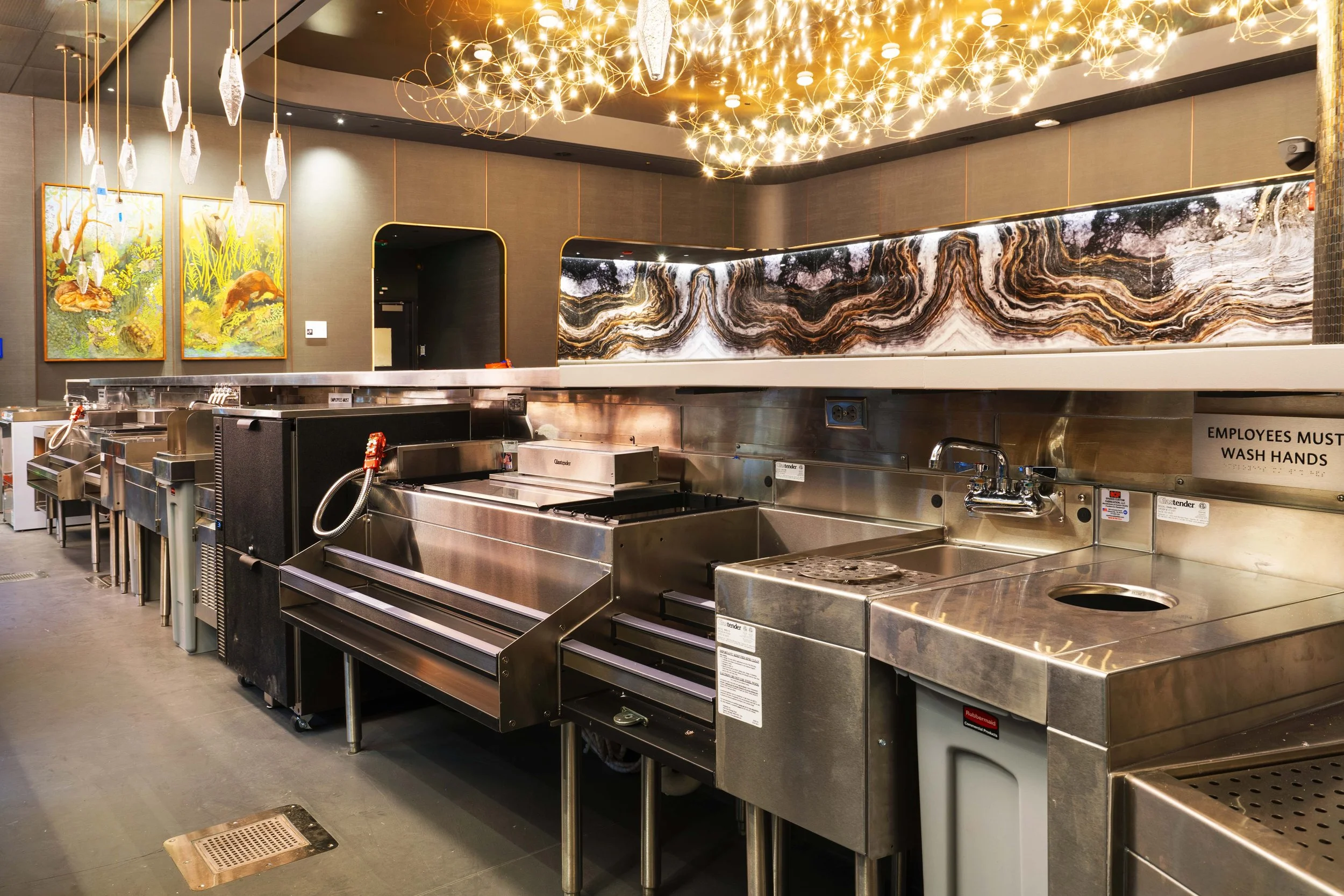 Empty commercial kitchen with stainless steel appliances, a sink, and decorative artwork on the wall, illuminated by hanging light fixtures.