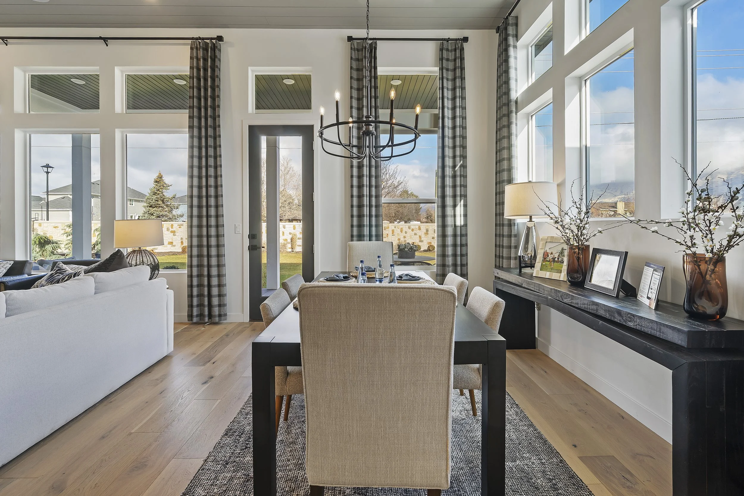 Living room with large windows, beige dining chairs around a black table, and a black chandelier hanging from the ceiling.