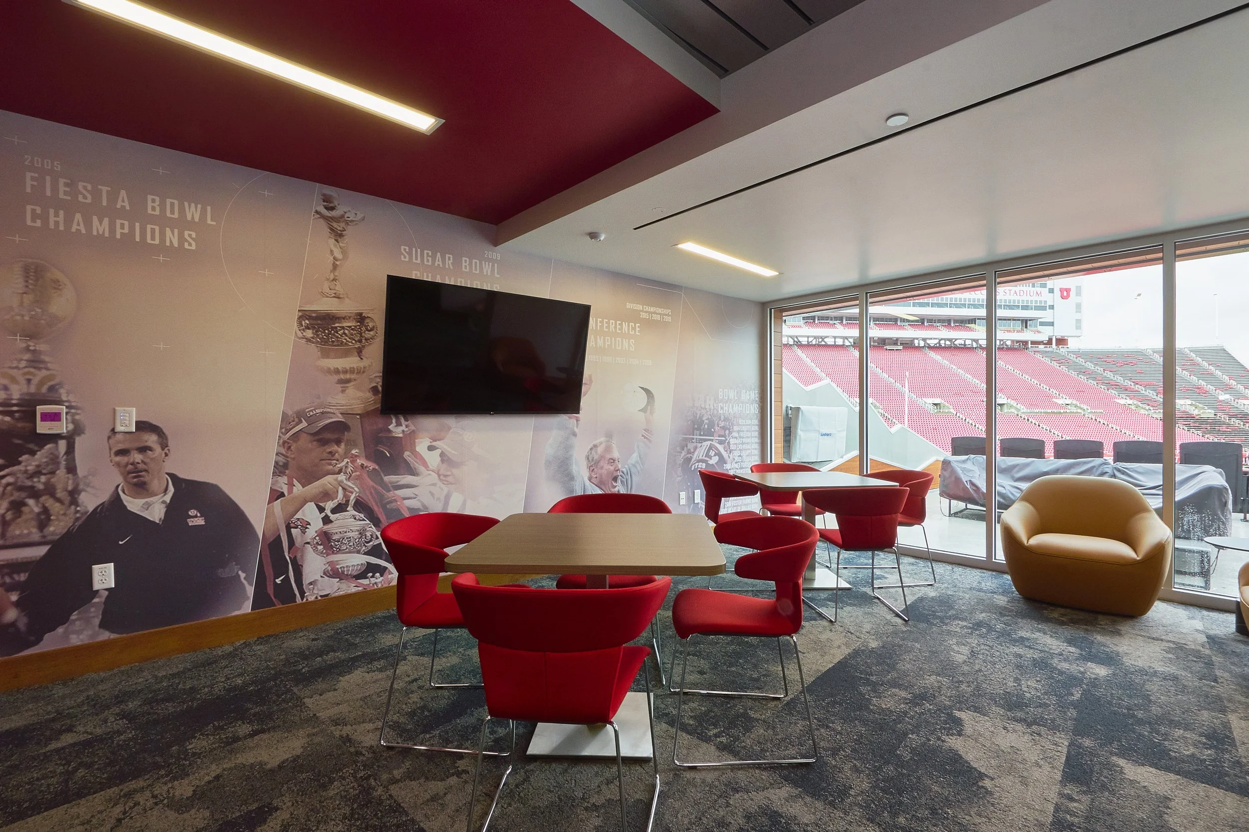 A lounge area inside a sports stadium with red chairs around a wooden table, a large beige armchair, and floor-to-ceiling windows overlooking a football field with seating and stadium structures.