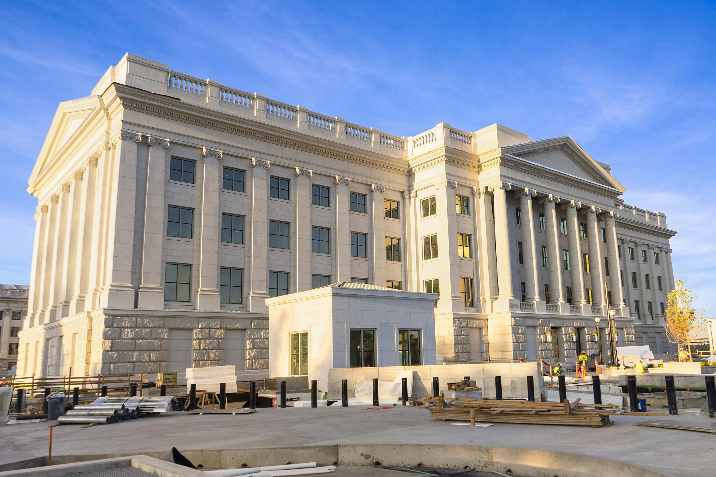 A large, classical style building with tall columns and multiple windows, under construction with construction materials and workers in front, against a blue sky.