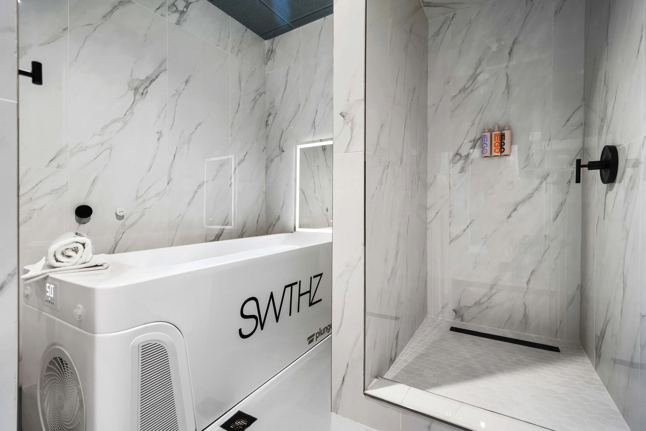 Modern bathroom with marble walls and floors, featuring a white bathtub with a rolled towel on top and a walk-in shower with black fixtures and four bottles of products.