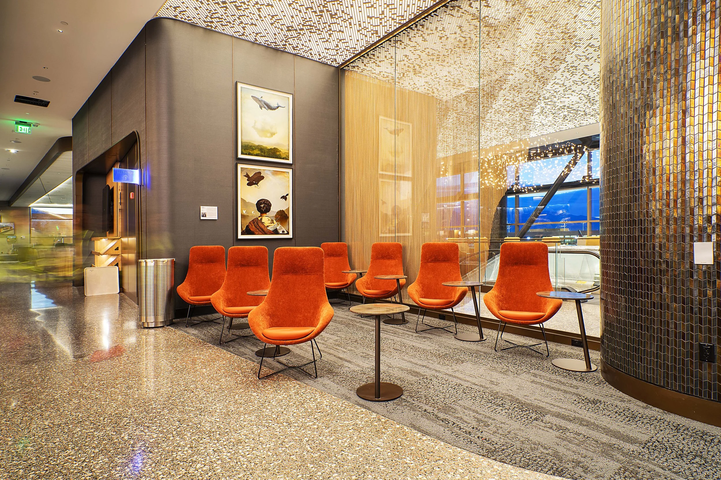 A modern airport lounge with orange chairs arranged in a semi-circle around small round tables, large windows with a view outside, colorful artwork of flying birds on the wall, and decorative ceiling lights and wall tiling.