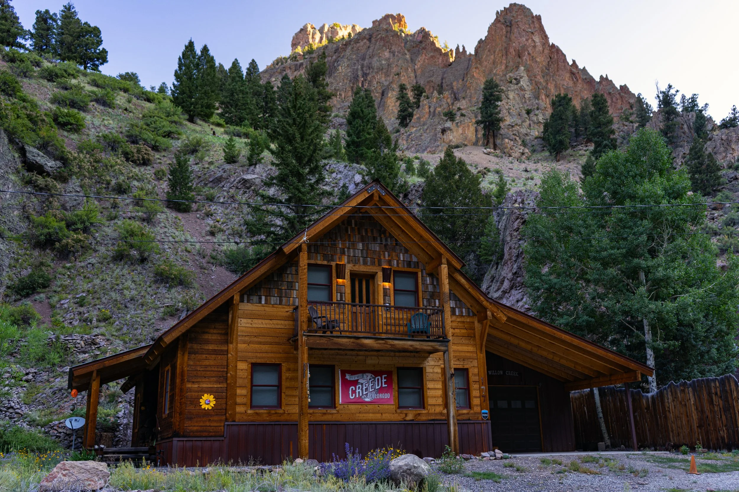 A rustic wooden cabin with a small balcony and a banner reading "Creede, Colorado," nestled in a mountainous area with surrounding pine trees and rock formations.