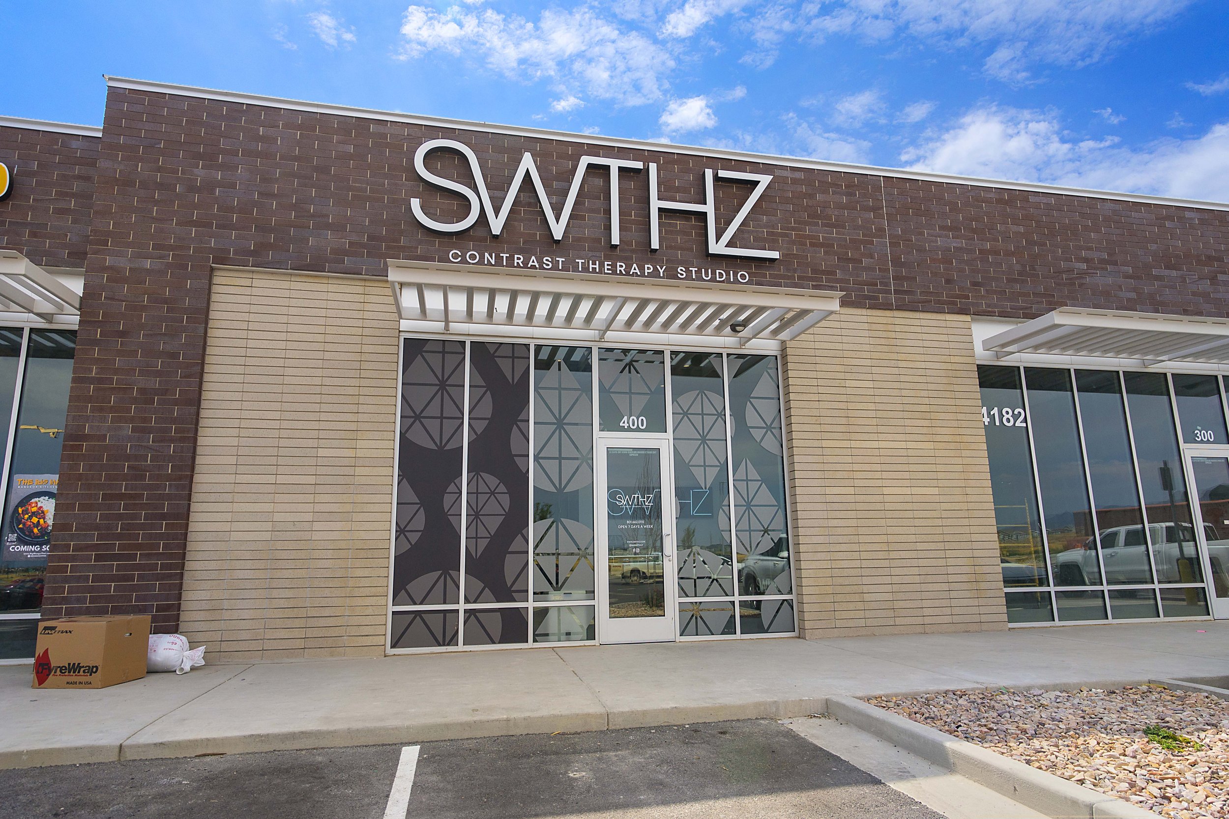 Exterior view of a commercial building with signage that reads 'SWTHZ Contrast Therapy Studio' in large letters, glass doors, and windows, with a parking lot in front and a partly cloudy sky above.