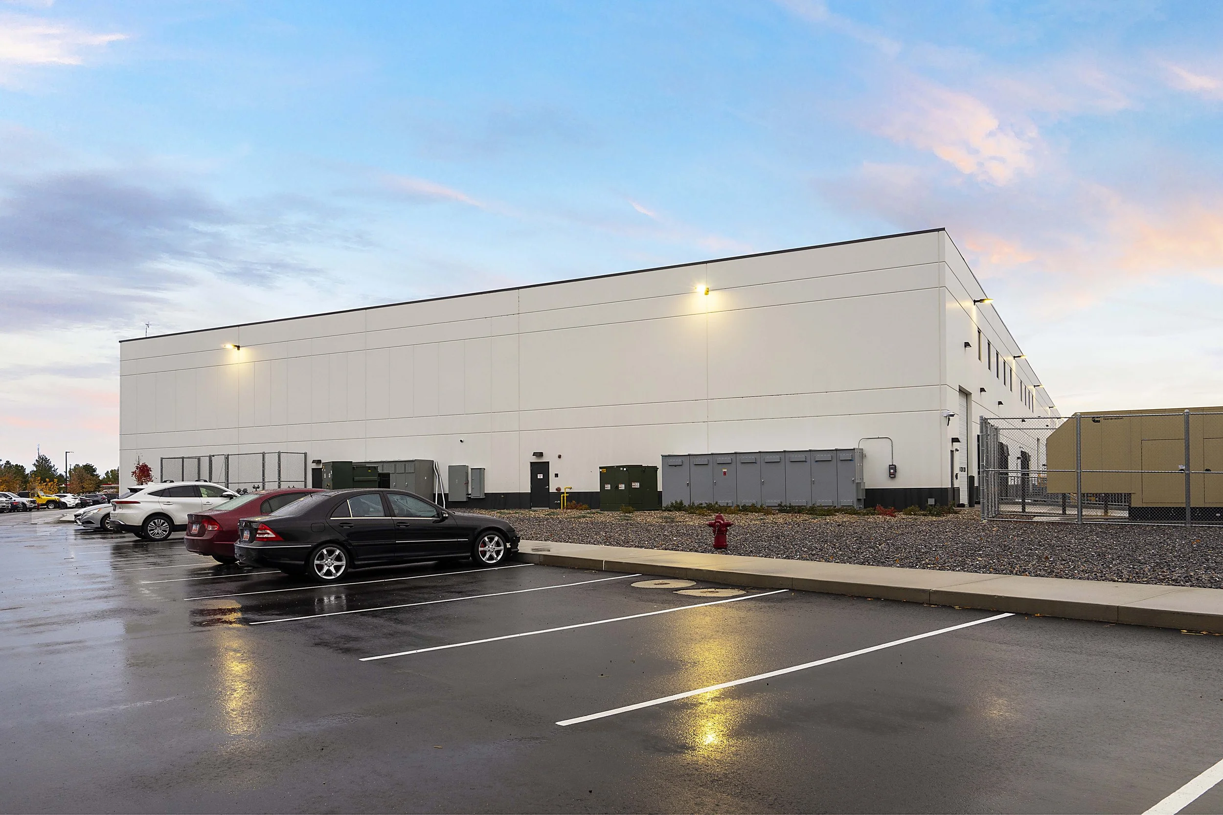 A parking lot after rain with several parked cars in front of a large white commercial building, under a partly cloudy sky at dusk.