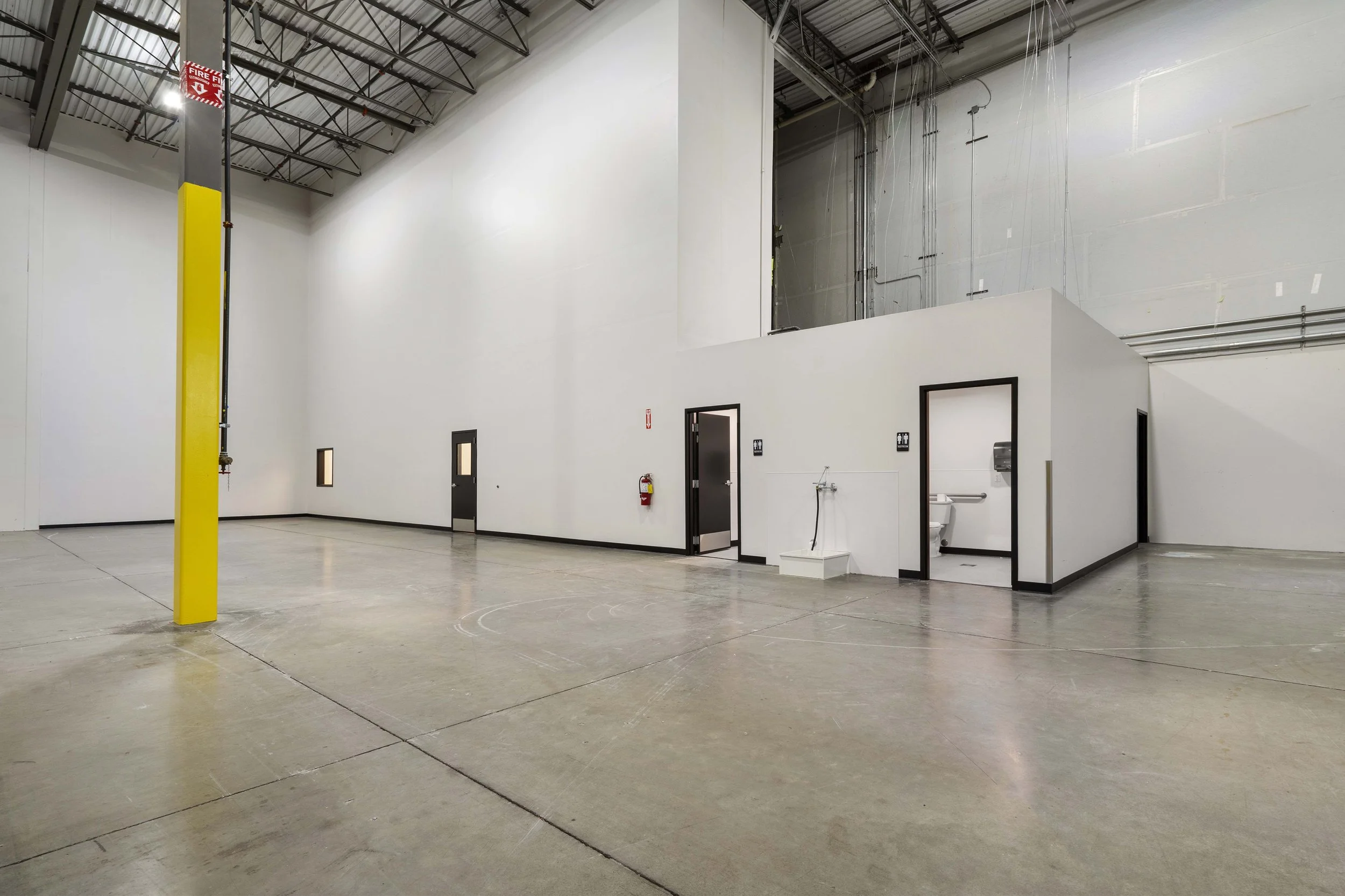 Empty warehouse or industrial space with a small enclosed restroom area, fire extinguisher, and yellow pole for fire safety, high ceiling with metal beams.