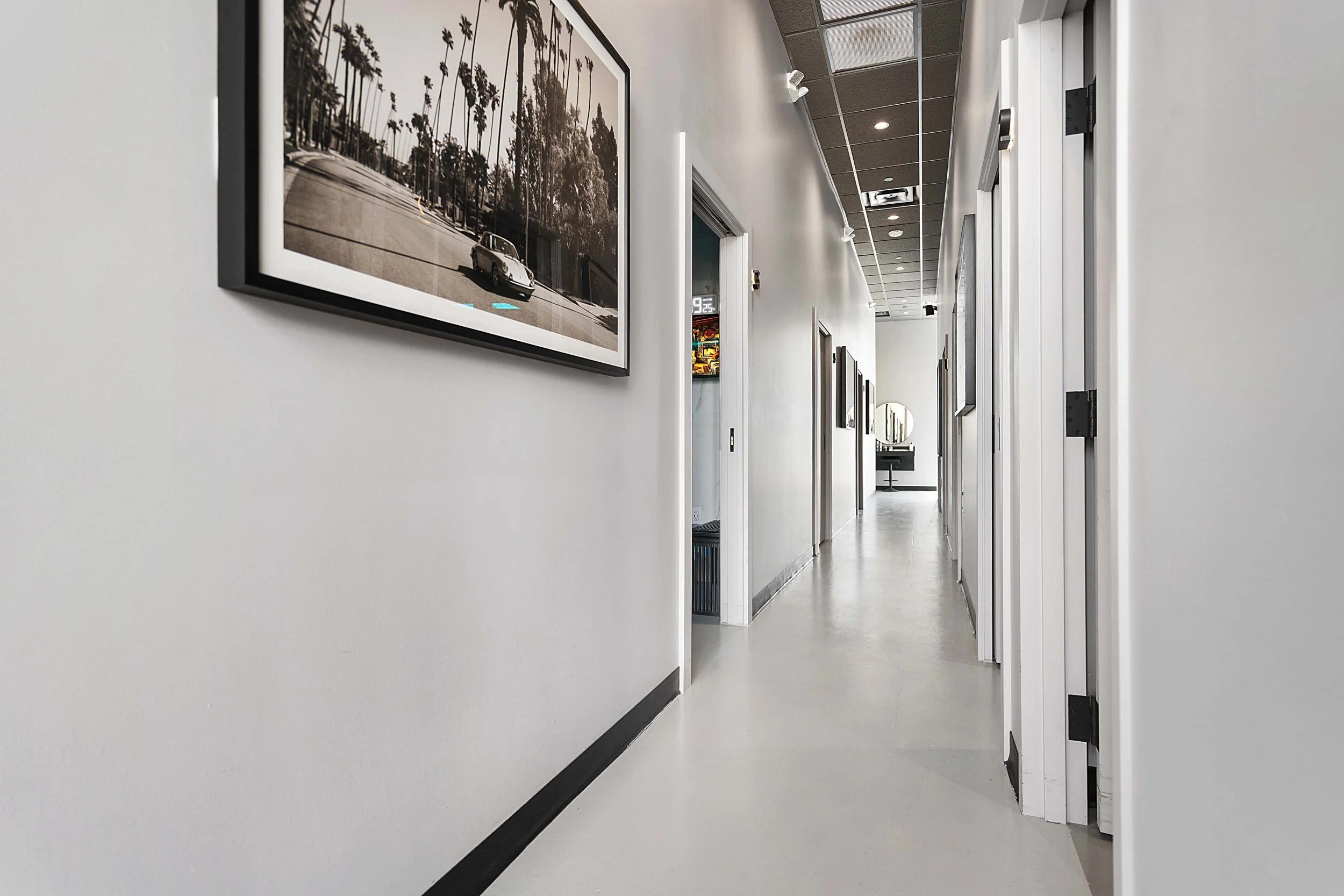 A long, narrow hallway in a modern building with white walls and a white floor. There are framed black-and-white photographs on the walls, and a mirror with a black frame at the end of the hallway. There are multiple doors along the sides of the hall