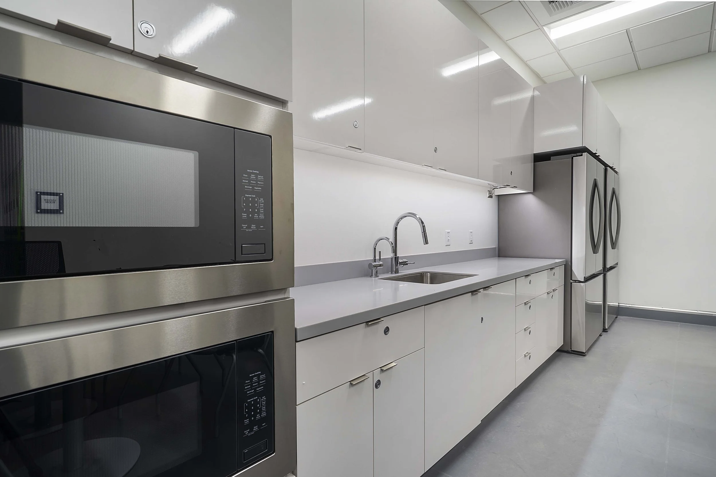 A clean, modern kitchen with white cabinets, stainless steel appliances including a microwave and refrigerator, a sink with a chrome faucet, and a gray floor.
