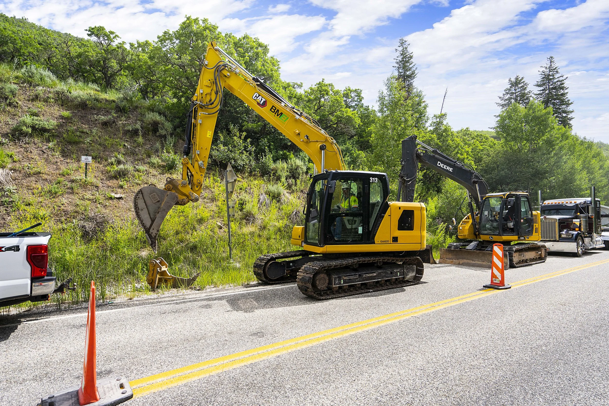 Construction workers operating excavators next to a highway, with traffic cones and parked trucks, surrounded by green trees and a partly cloudy sky.