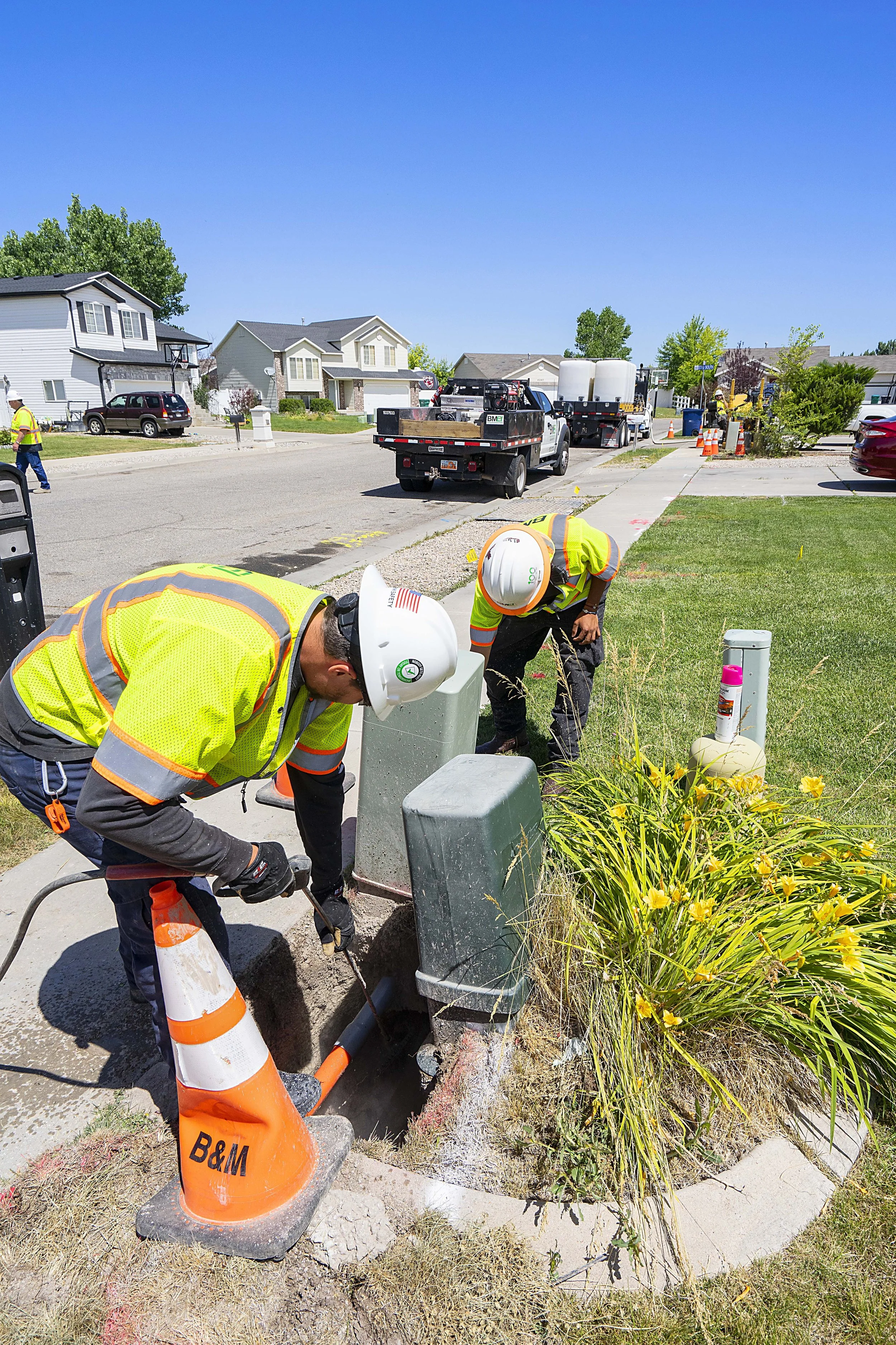 Workers in high-visibility safety gear repairing underground equipment on a sidewalk with utility boxes and plants, with a residential street and parked vehicles in the background.