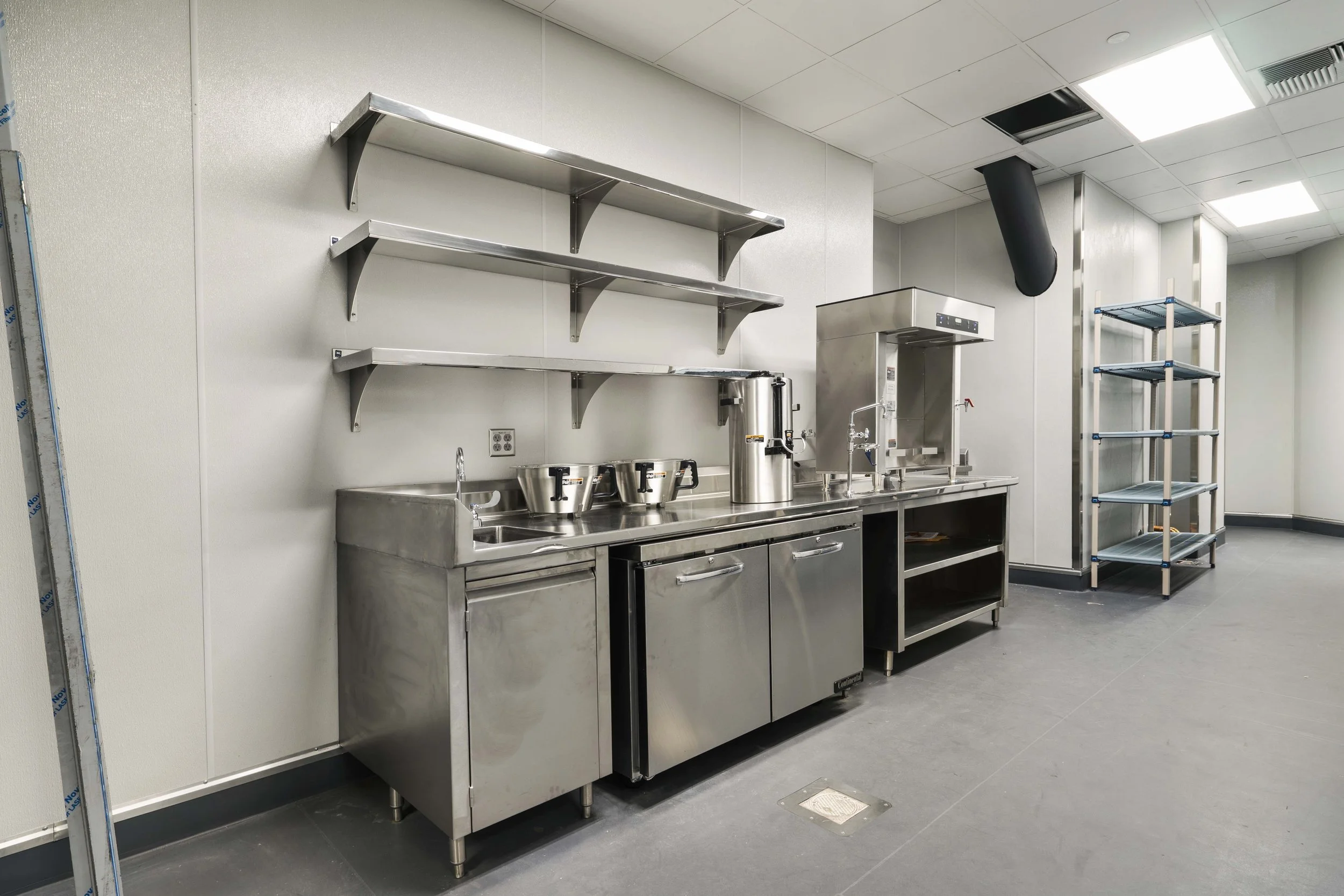 A commercial kitchen with stainless steel counters, sink, shelving, and a water boiler, set against white walls and a gray floor.