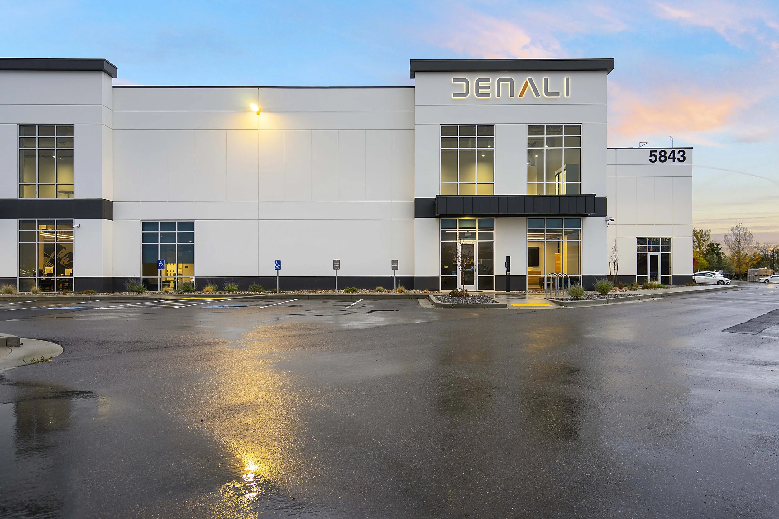 Exterior view of a modern commercial building with the number 5843 on the wall, large windows, and a parking lot with a few cars and wet pavement reflecting the building and sunset sky.