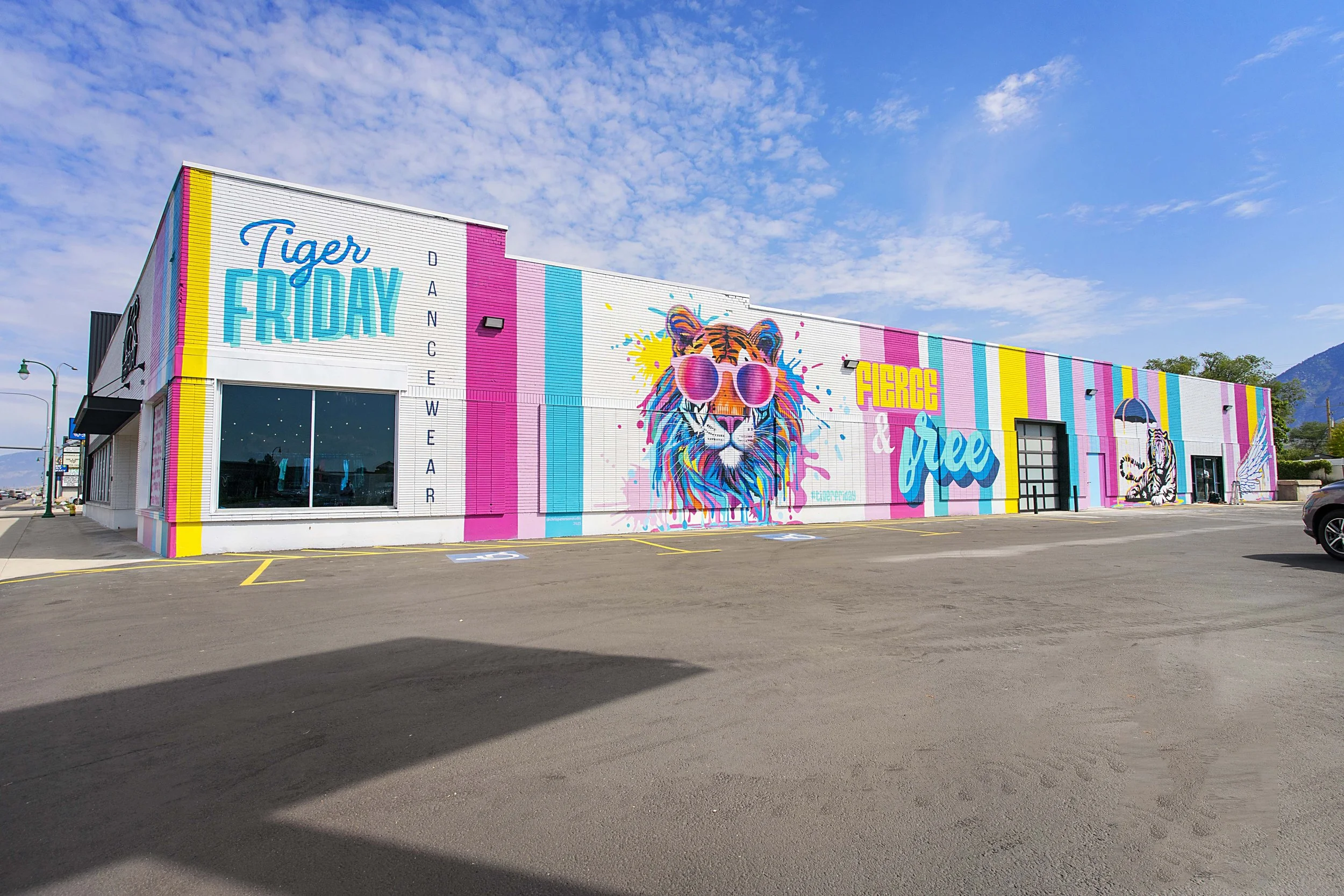Colorful mural on the side of a building featuring a lion wearing pink sunglasses and a white tiger, with the words 'Tiger Friday' and 'Dancewear' on the wall.